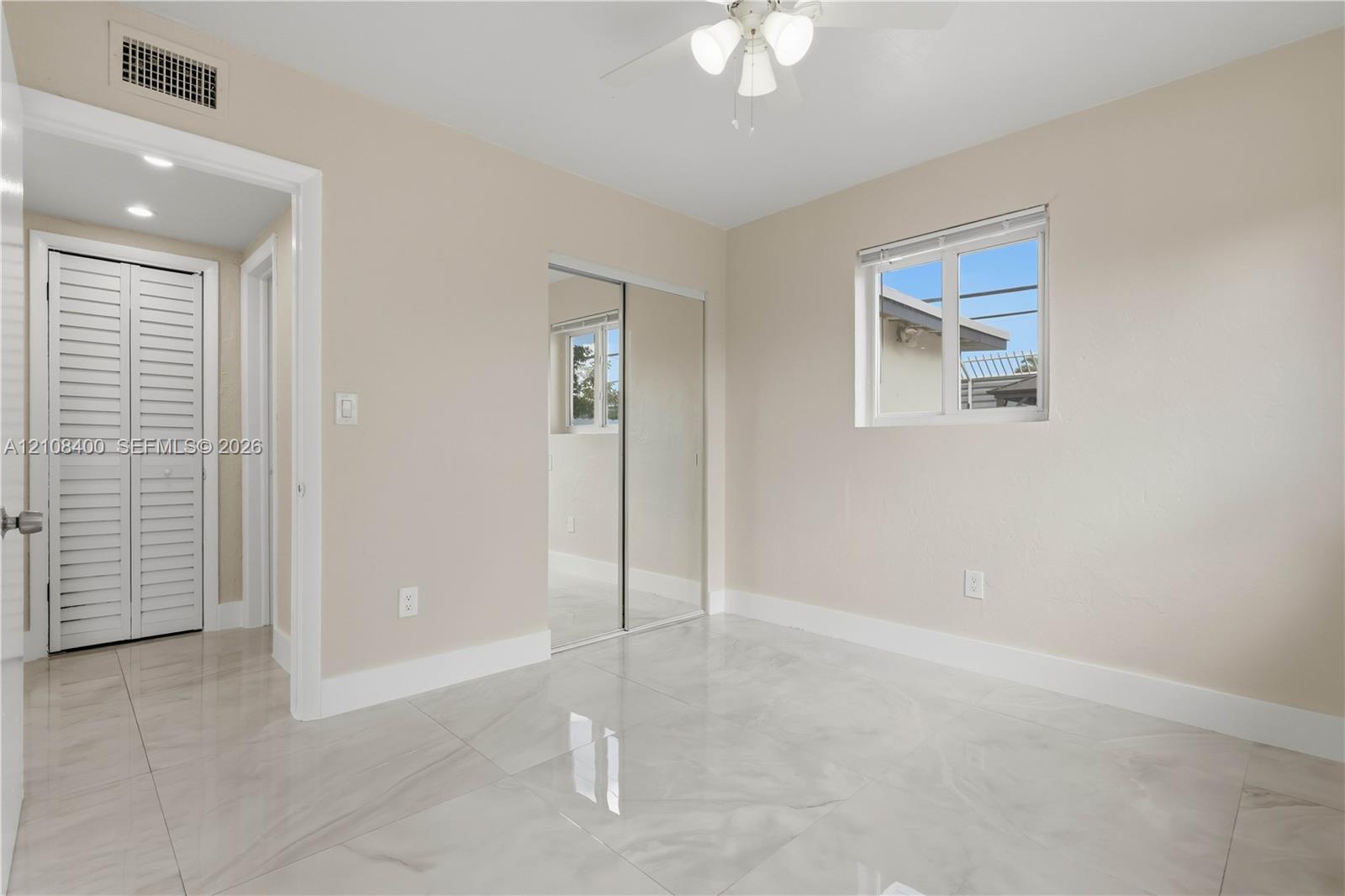 Neutral-toned room featuring large-format tile flooring, mirrored sliding-door closet, multi-pane window, and integrated ceiling fan