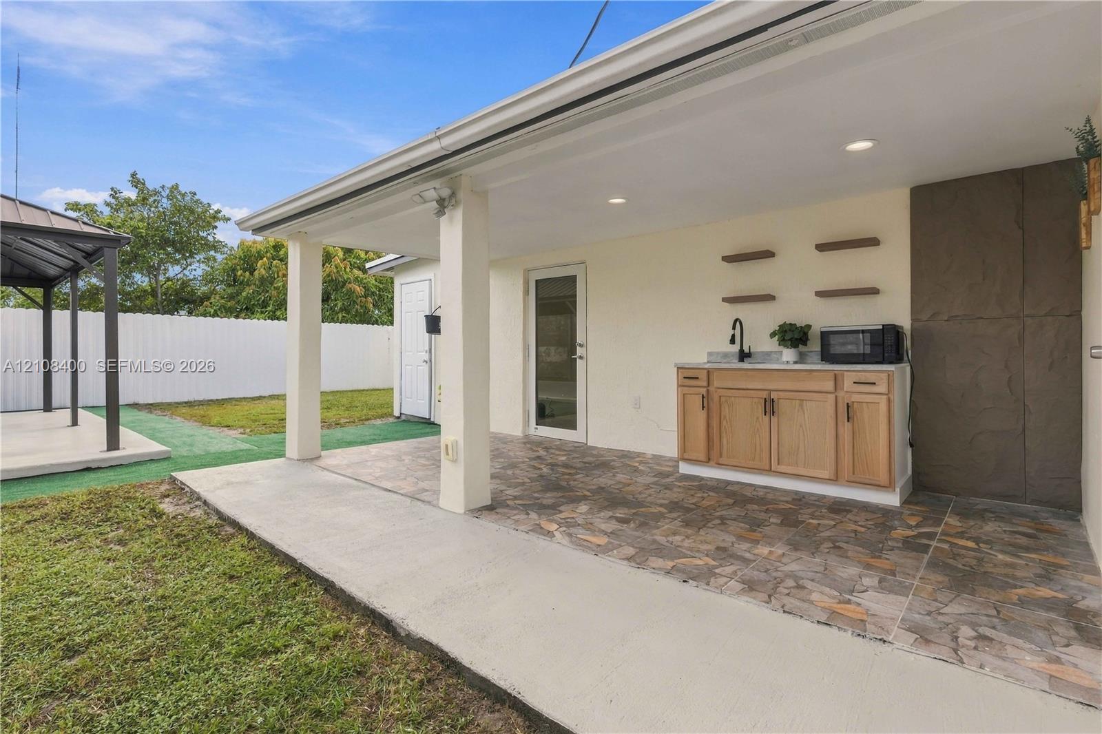 Covered patio featuring stone-tile flooring, recessed lighting, and an outdoor kitchenette with light wood-finish cabinetry, a stone countertop, and a matte black faucet