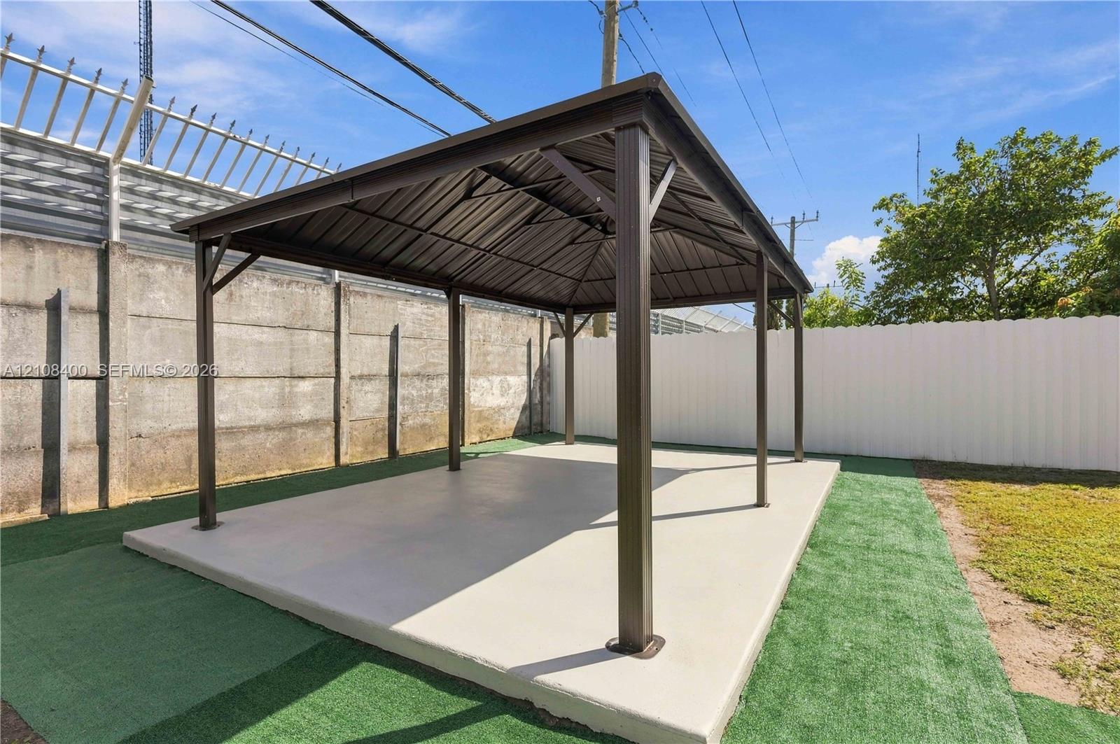 Outdoor gazebo featuring a dark metal frame, a peaked metal roof, and a concrete slab foundation, bordered by a concrete perimeter wall and synthetic turf