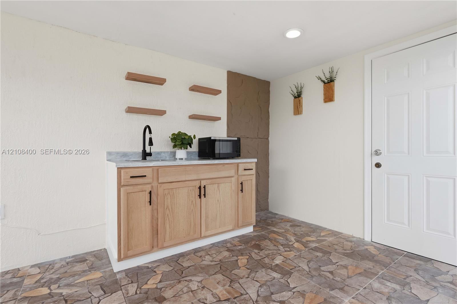 Custom cabinetry with light wood-finish doors, stone-pattern countertop, matte black faucet, floating wall shelving, and flagstone-style flooring