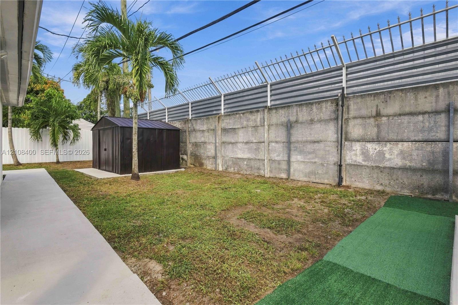 Backyard featuring concrete slab surfacing, a metal storage shed, synthetic turf sections, and a concrete perimeter wall with an integrated metal fence topper