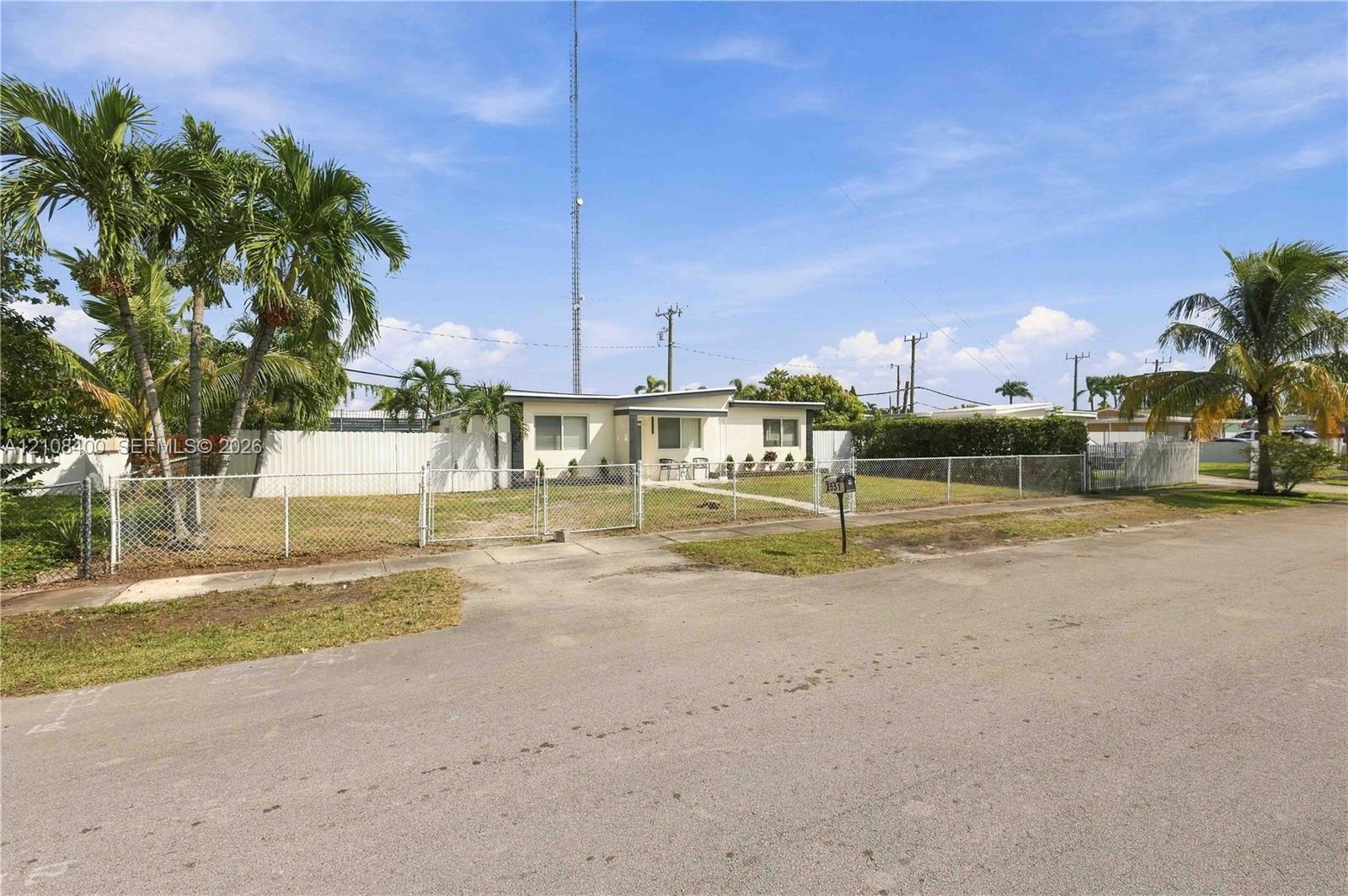 Single-story exterior featuring a stucco facade, chain-link fencing, mature palm trees, and an asphalt driveway