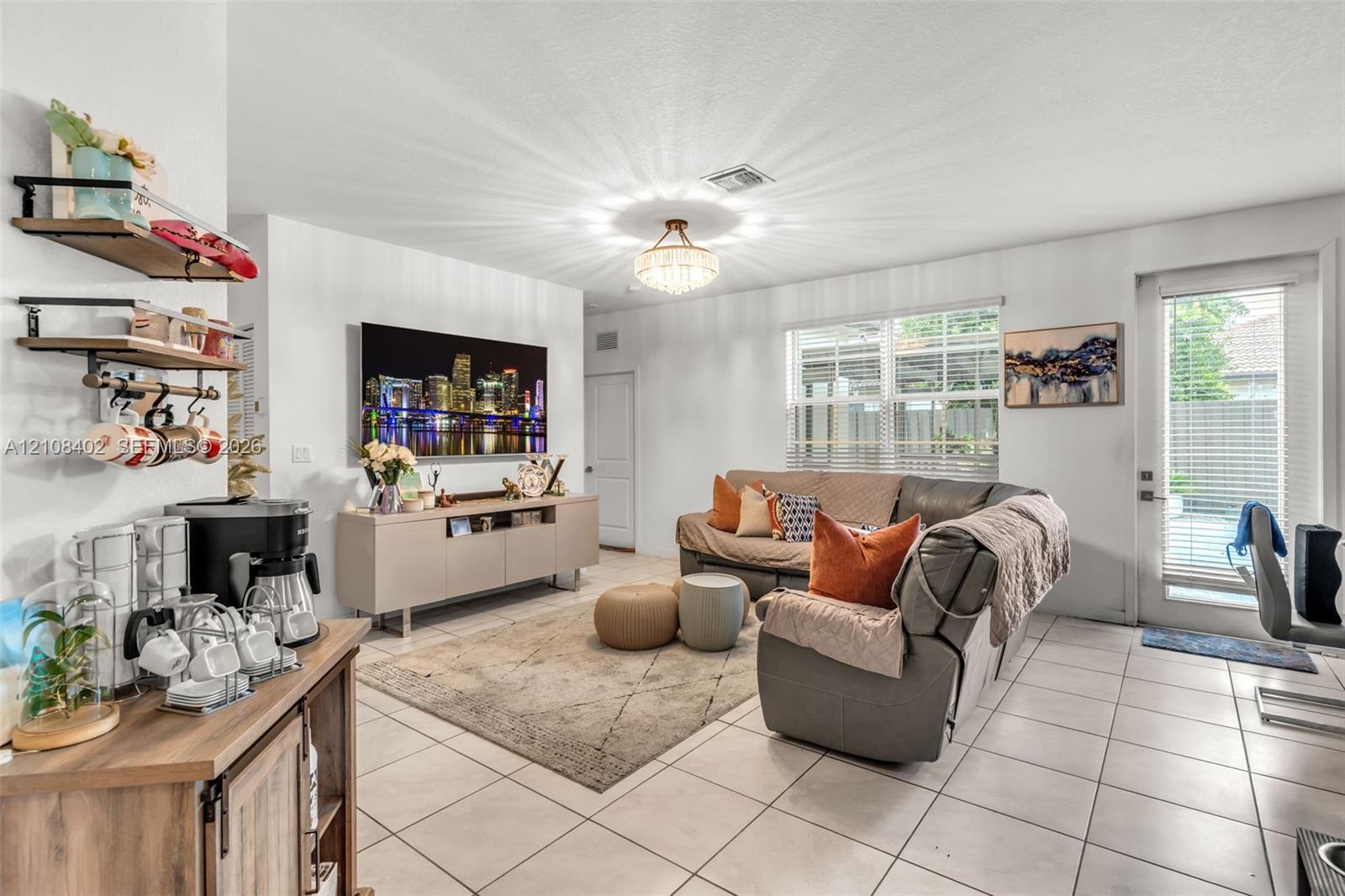 Living area featuring tile flooring, a multi-tiered crystal-style chandelier, horizontal window blinds, and a glass-paneled exterior door