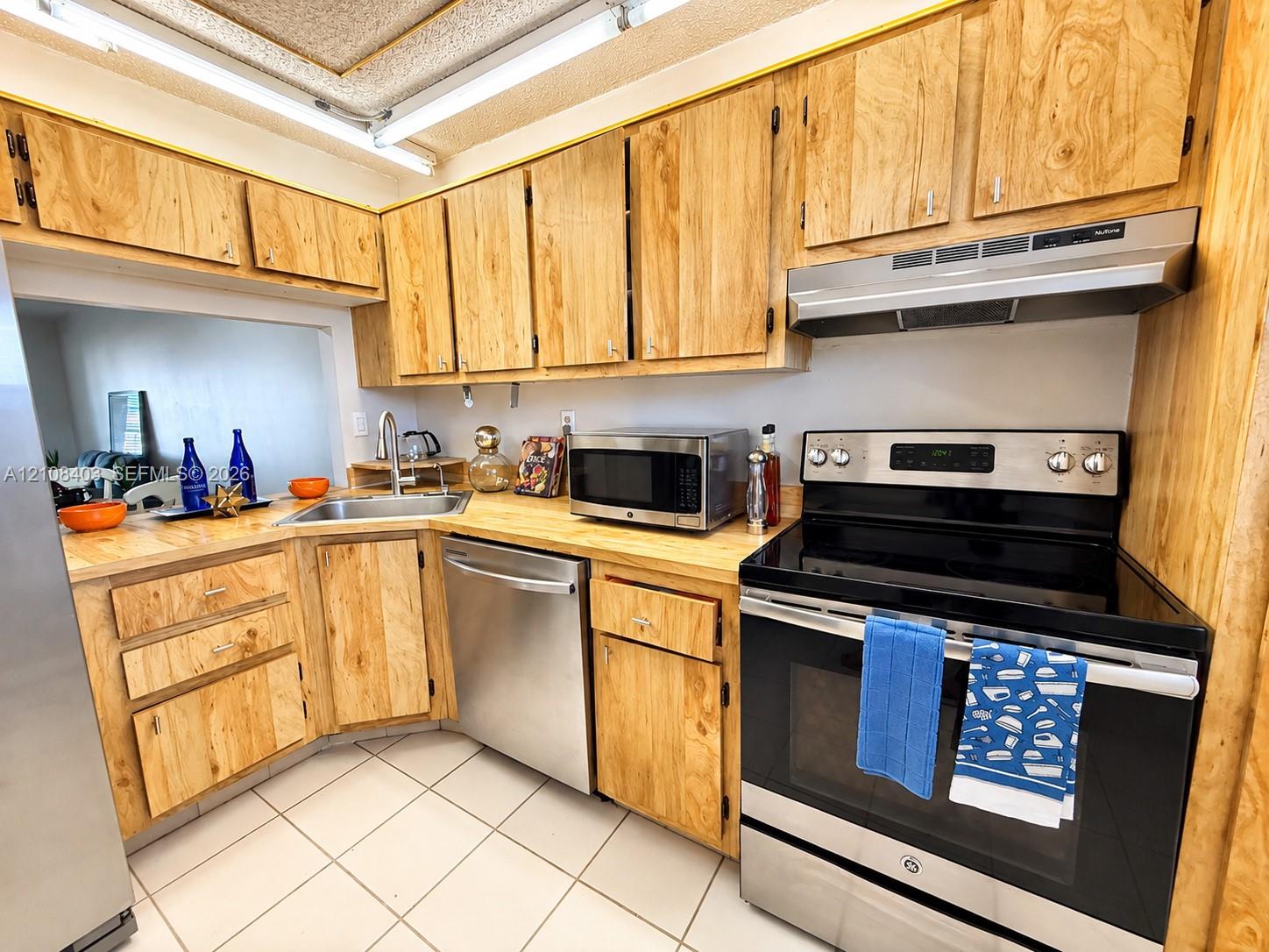 Kitchen featuring wood cabinetry, stainless steel appliances, neutral tiled flooring, and a countertop mounted sink Kitchen featuring wood cabinetry, stainless steel appliances, neutral tiled flooring, and a countertop mounted sink