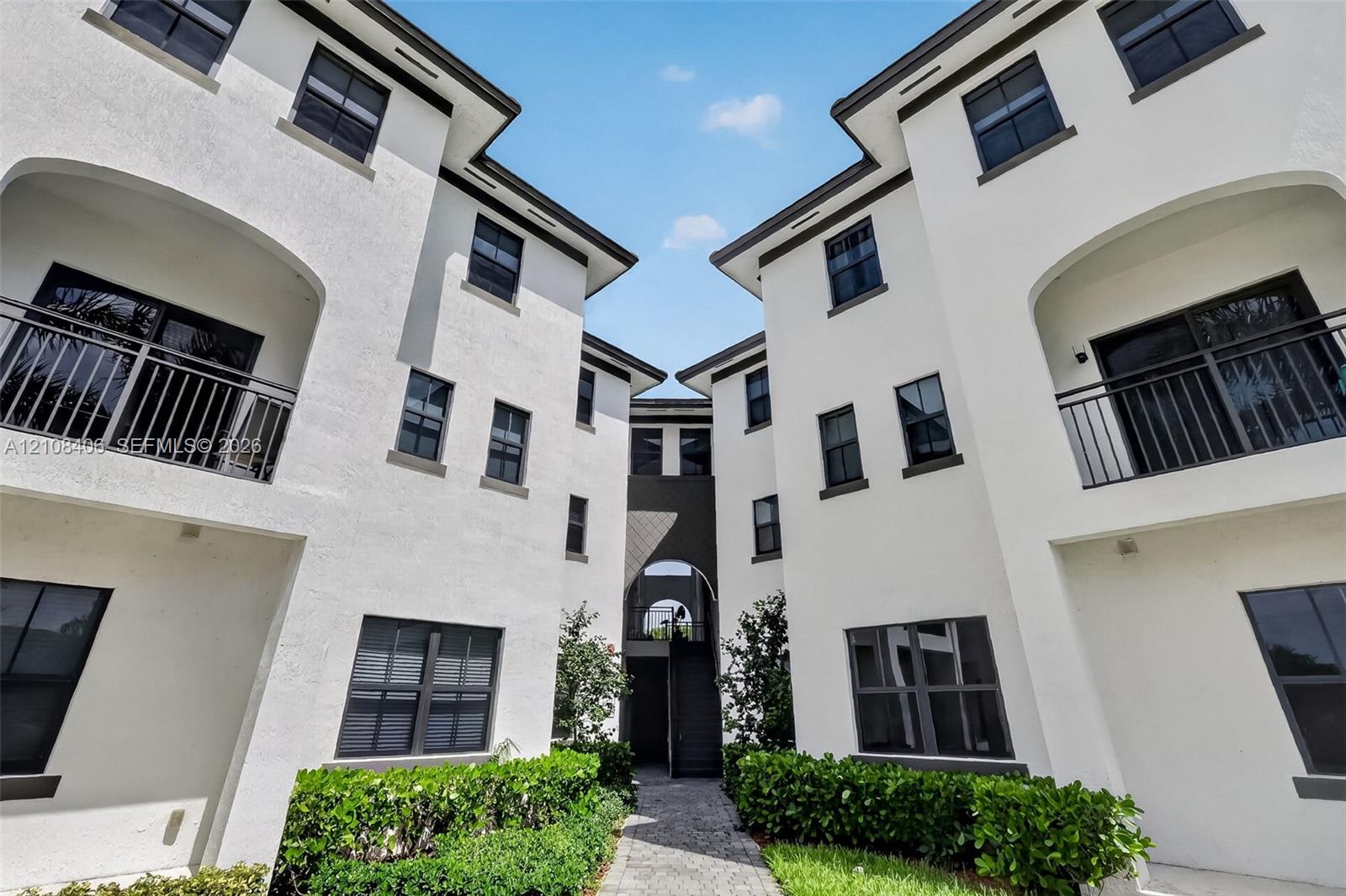 Multi-story stucco exterior featuring arched balcony detailing, dark-framed windows, paved walkways, and manicured shrubbery