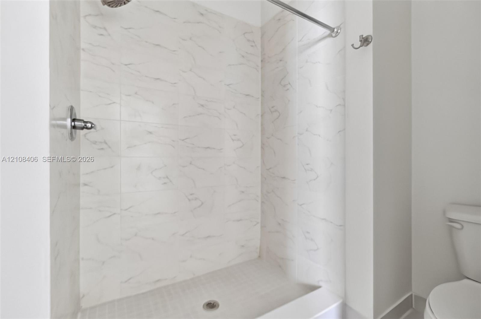 Shower enclosure featuring white tile with grey veining, a wall-mounted shower head, a chrome shower valve, a metal shower curtain rod, and a wall-mounted hook
