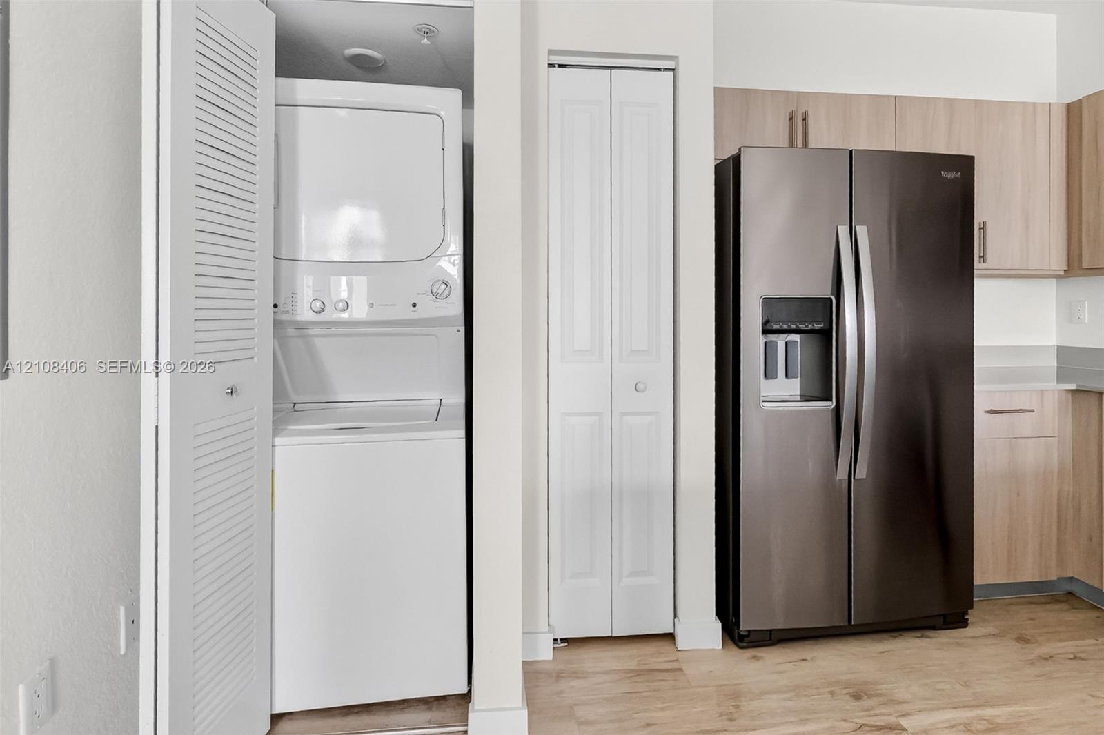 Built-in laundry closet with stacked washer and dryer, side-by-side stainless steel refrigerator, light-tone wood-finish cabinetry, and wood-finish flooring