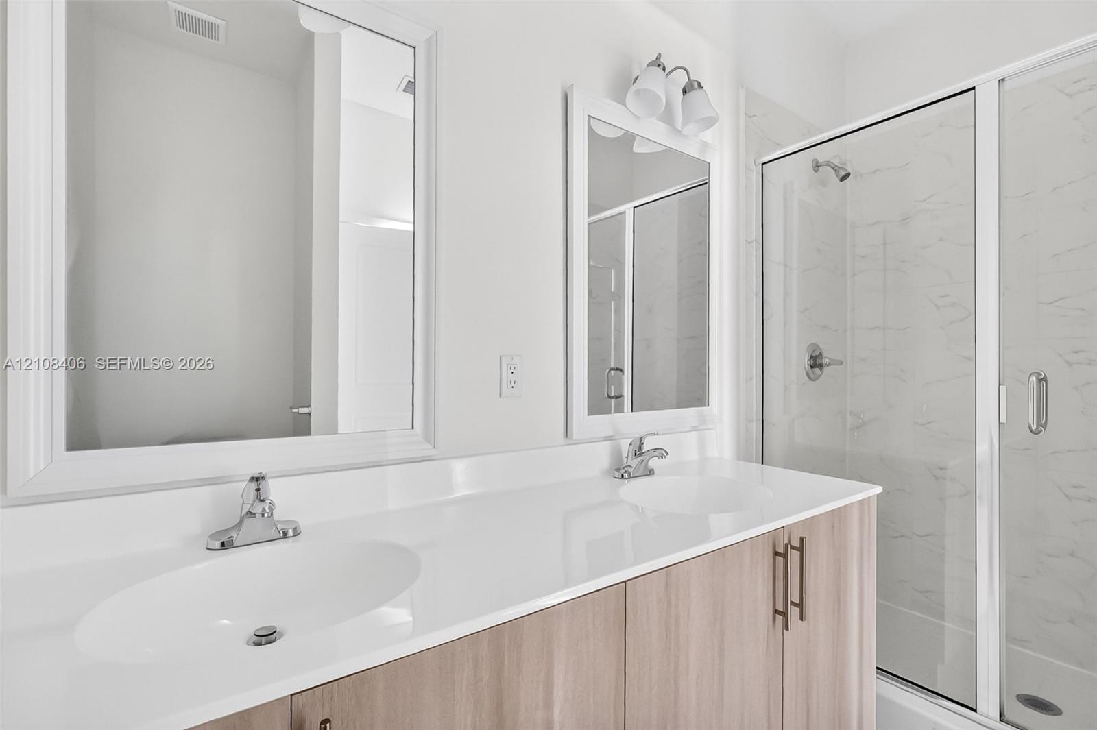 Dual-sink vanity with a white countertop and wood-finish cabinetry, framed mirrors, and a glass-enclosed shower with light-toned wall tile