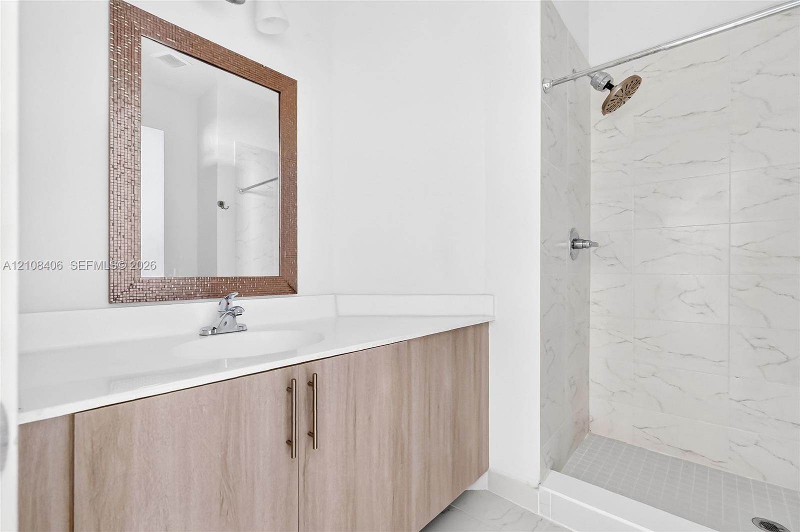 Bathroom vanity featuring a wood-finish cabinet, white solid-surface countertop, and rectangular mirror with a mosaic frame, alongside a walk-in shower with light-toned wall tile and a silver-finish shower head