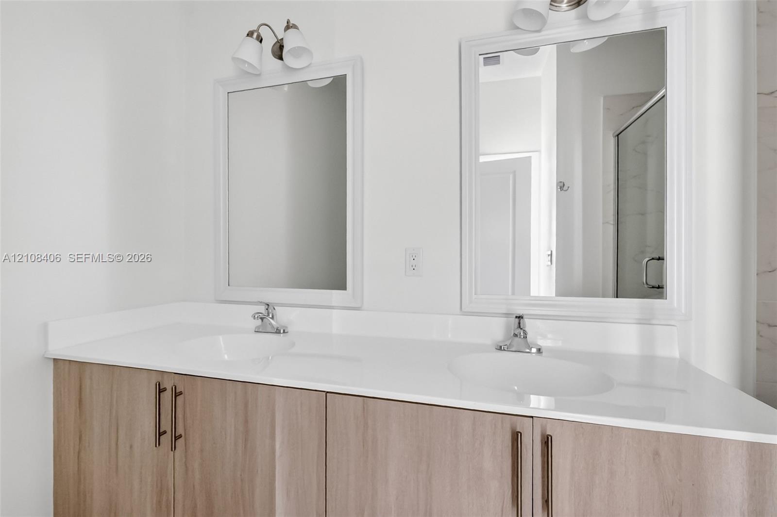 Dual-sink vanity with a white solid-surface countertop, light wood-finish cabinetry, brushed metal hardware, and framed mirrors with overhead light fixtures