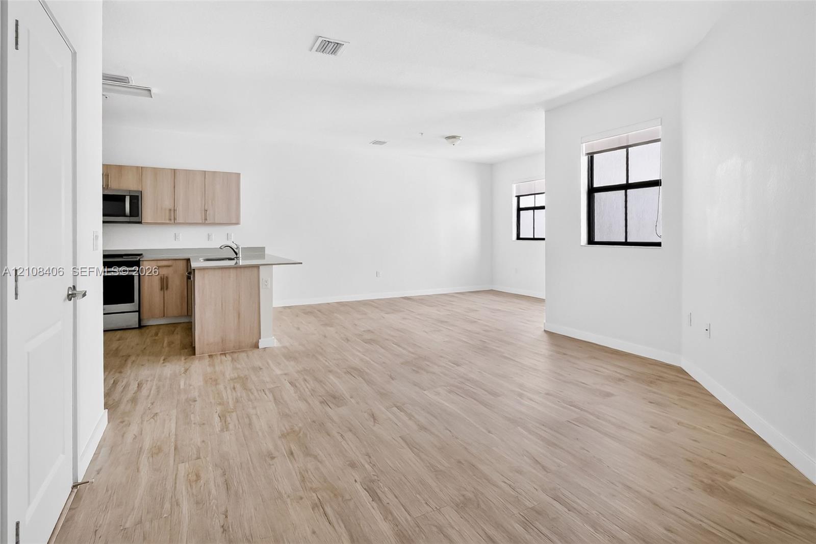 Open-concept floor plan featuring wood-finish flooring, cabinetry with a light natural finish, stainless steel appliances, and large windows with black frames