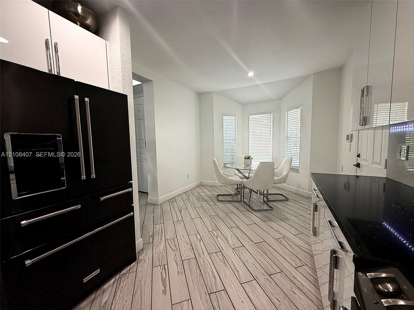 Modern kitchen featuring white high-gloss cabinetry, black polished countertops, a professional-style black refrigerator, and wood-finish flooring with a bay window configuration Modern kitchen featuring white high-gloss cabinetry, black polished countertops, a professional-style black refrigerator, and wood-finish flooring with a bay window configuration