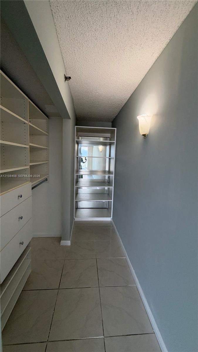 Walk-in closet featuring white built-in shelving, integrated storage drawers with silver-tone hardware, tile flooring, and a wall-mounted sconce
