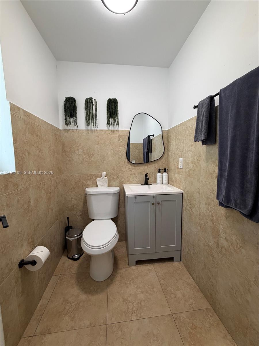 Neutral-toned wall and floor tiling, gray vanity cabinet with a white countertop, organic-shaped wall mirror, and matte black hardware