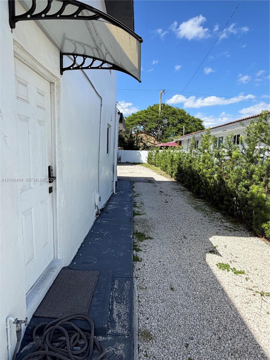 Exterior side entrance featuring a curved metal door awning, a painted concrete walkway, and a gravel driveway bordered by a privacy hedge