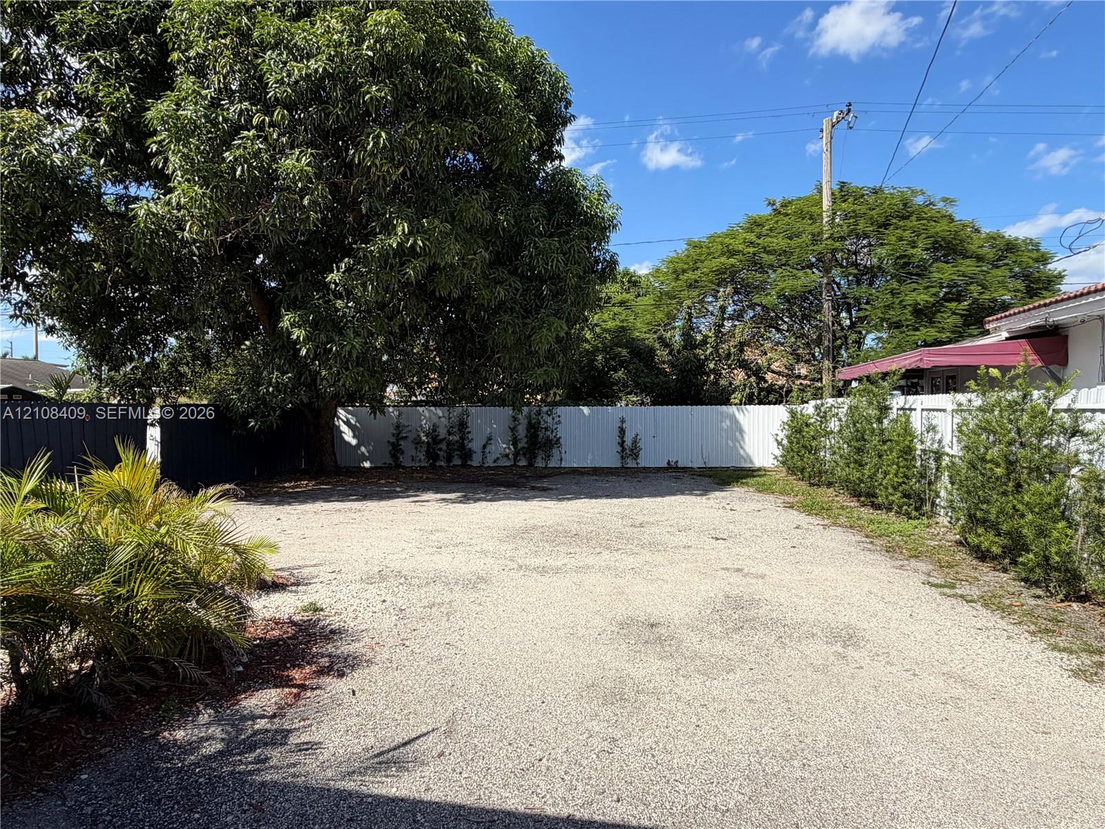 Gravel-surfaced outdoor area enclosed by white vinyl fencing and lined with mature trees