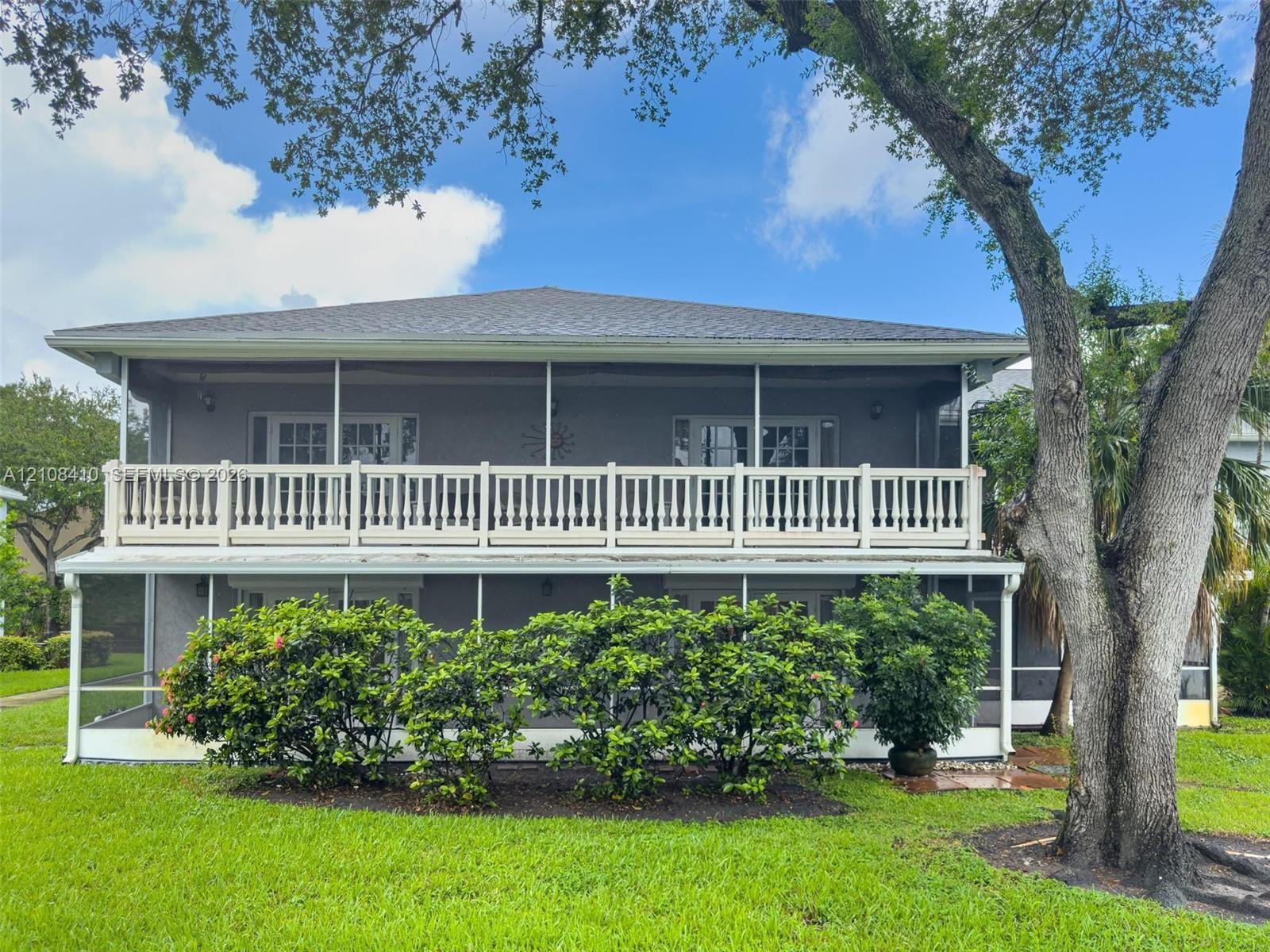 Two-story exterior featuring a screened-in ground level, an upper-level balcony with white balustrade railings, and mature trees