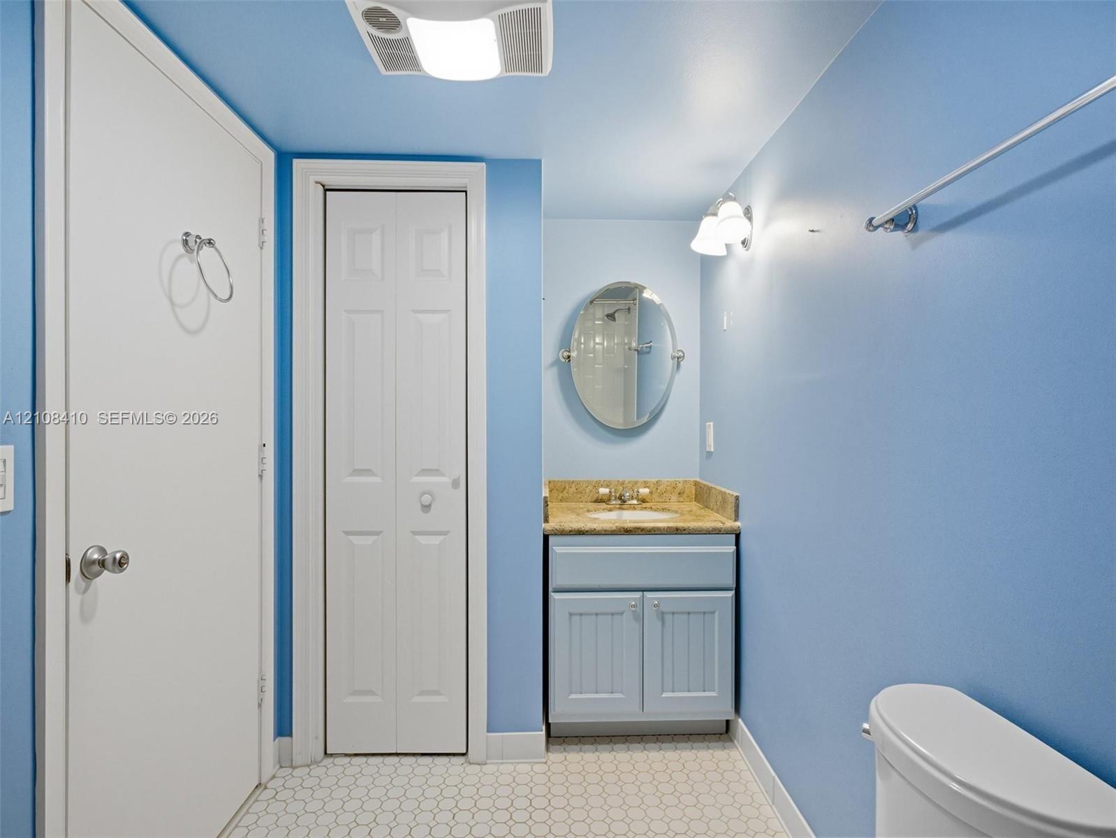 Bathroom featuring a vanity with stone-finish countertop, oval wall mirror, wall-mounted light fixture, bi-fold closet door, and hexagonal tile flooring