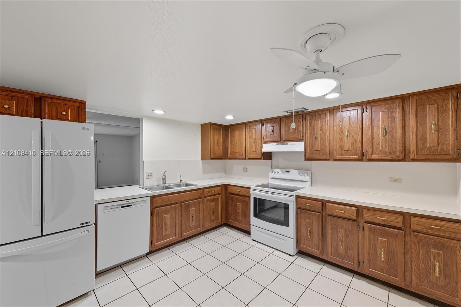 Kitchen featuring natural wood cabinetry, white appliances, a double-basin sink, white countertops, and tile flooring Kitchen featuring natural wood cabinetry, white appliances, a double-basin sink, white countertops, and tile flooring
