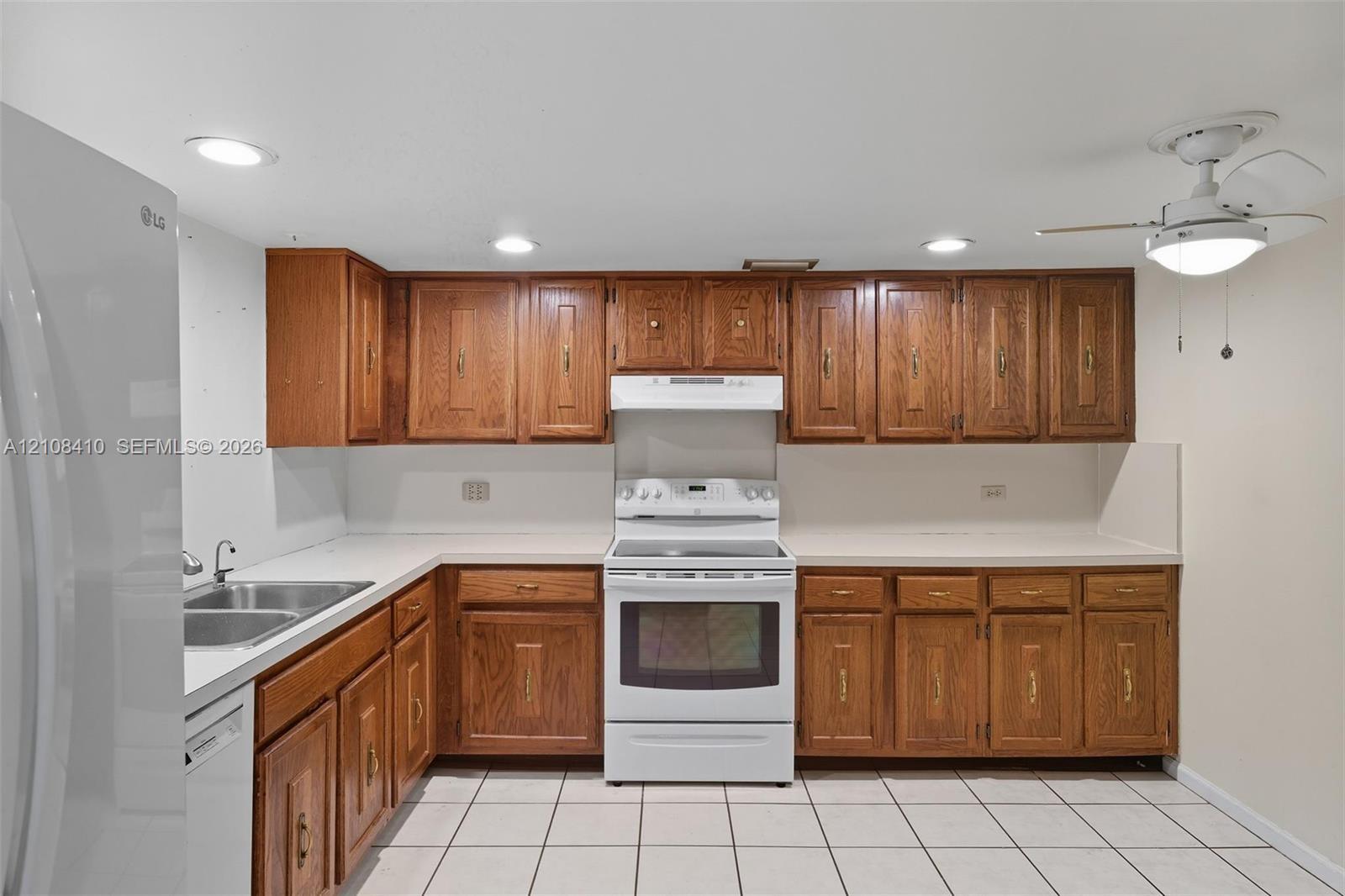 Kitchen featuring wood cabinetry, white appliances, a double-basin stainless steel sink, solid-surface countertops, and ceramic tile flooring Kitchen featuring wood cabinetry, white appliances, a double-basin stainless steel sink, solid-surface countertops, and ceramic tile flooring