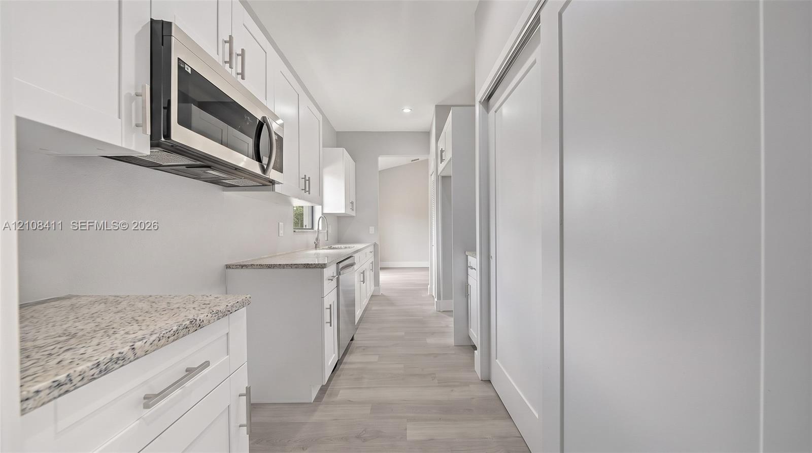 White shaker-style cabinetry featuring satin nickel hardware, speckled stone-finish countertops, stainless steel over-the-range microwave, built-in dishwasher, and wood-finish flooring