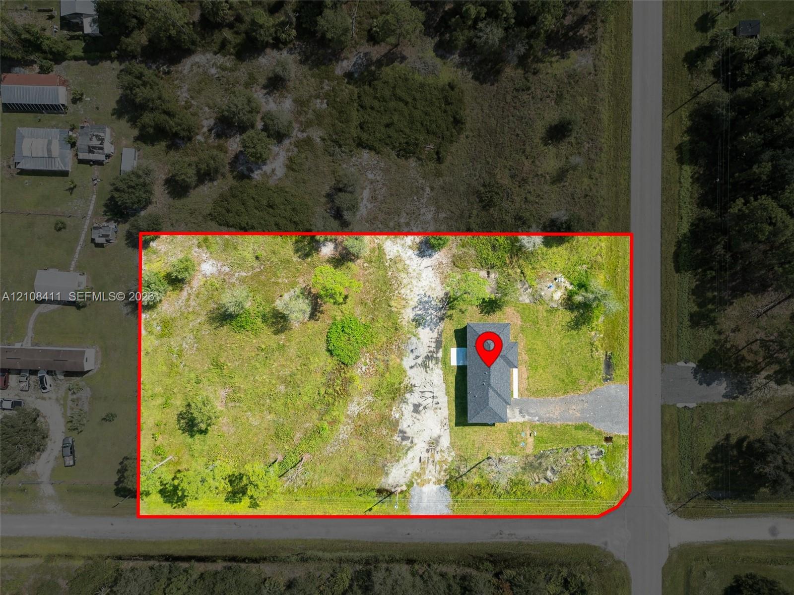 Aerial view of a corner lot featuring a detached structure, gravel driveway, mature trees, and expansive open green space
