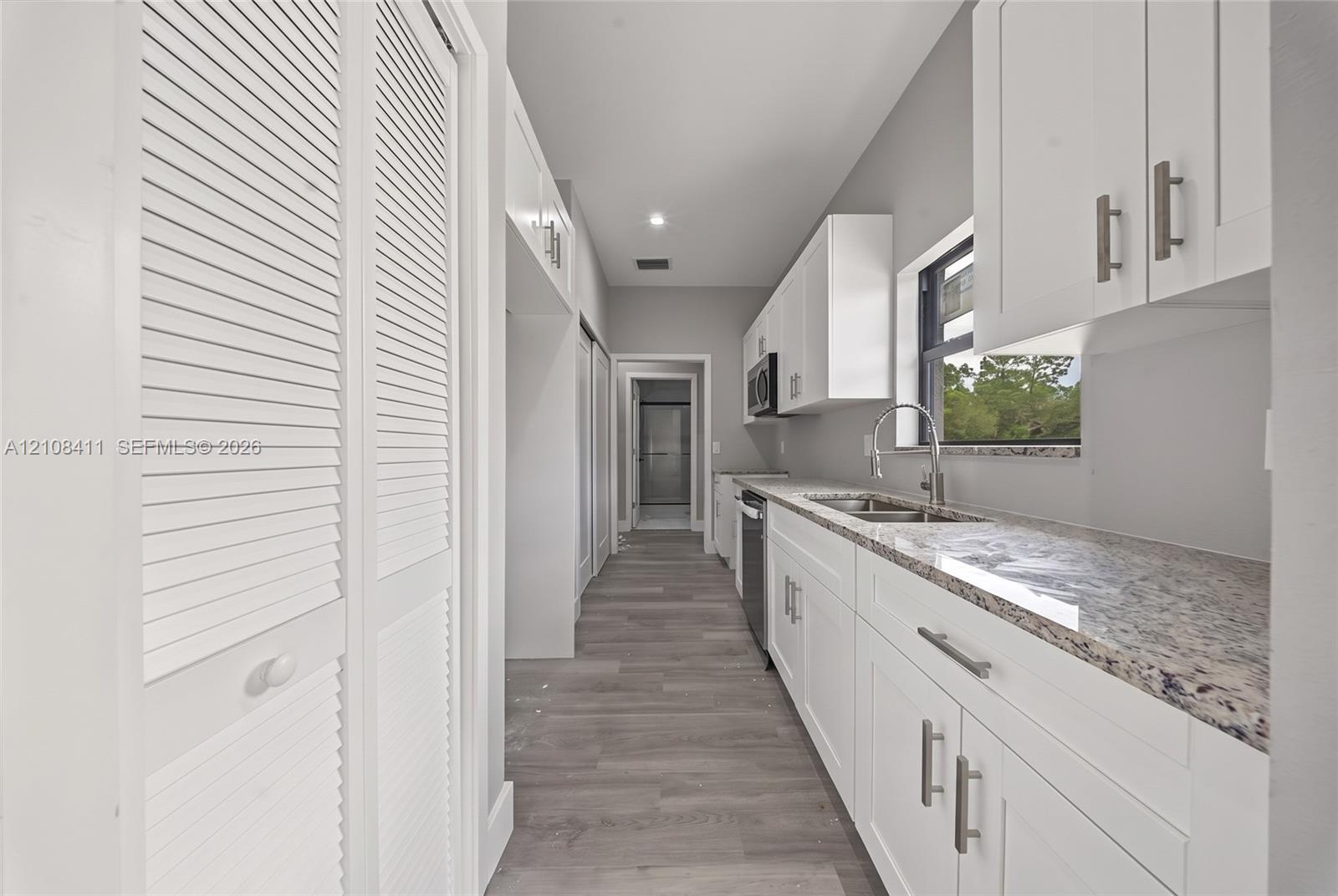Galley kitchen featuring white Shaker-style cabinetry, stone-finish countertops, stainless steel undermount sink with a gooseneck faucet, wood-finish flooring, and white louvered-door closets