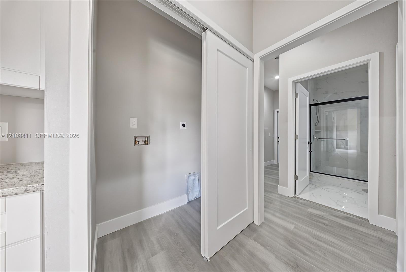 Utility space featuring light-toned wood-finish flooring, white cabinetry with speckled stone-finish countertops, and a sliding door, with a glimpse of a walk-in shower containing light-toned stone-finish tile and a black-framed glass enclosure