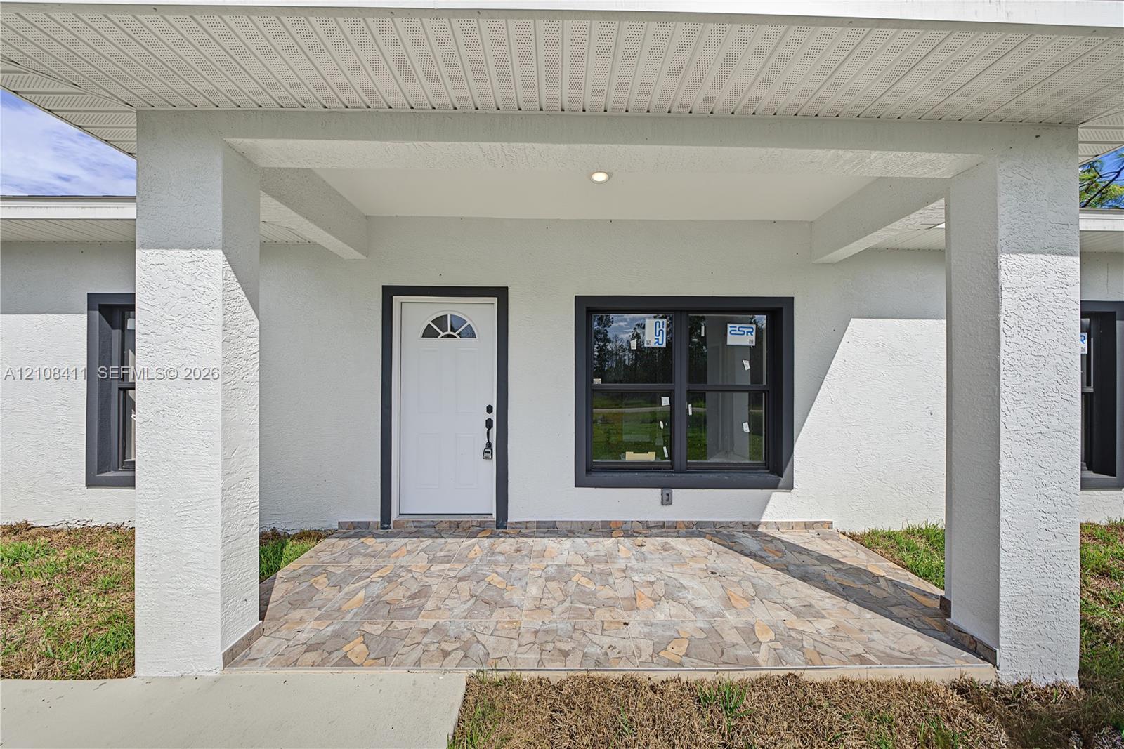 Covered entryway featuring a stone-paver porch, structural support columns, white door with a semi-circular transom window, and dark-framed windows Covered entryway featuring a stone-paver porch, structural support columns, white door with a semi-circular transom window, and dark-framed windows