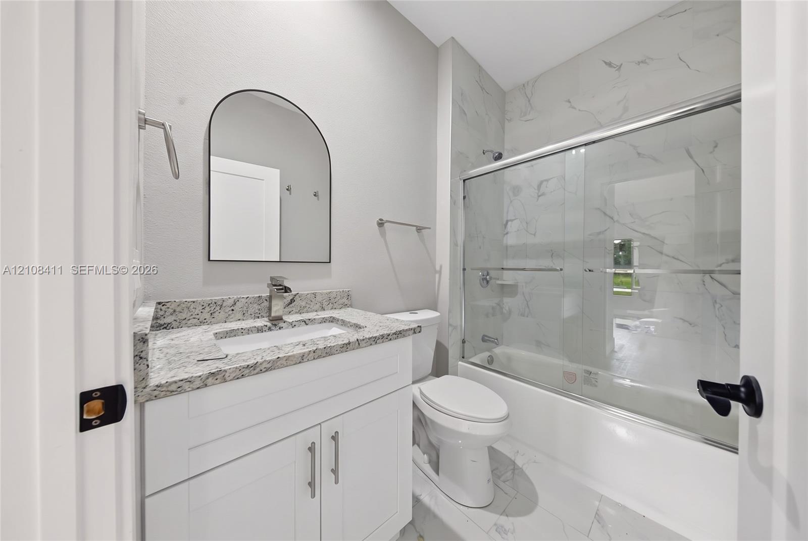 Bathroom featuring a white shaker-style vanity, stone-finish countertop, arched wall-mounted mirror, and a shower-tub combination with full-height tile surround