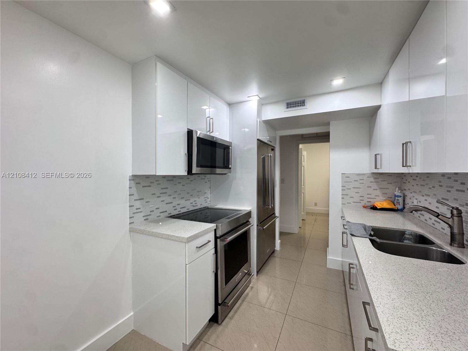 Kitchen featuring high-gloss white cabinetry, stone-finish countertops, stainless steel appliances, a mosaic tile backsplash, and large-format tile flooring