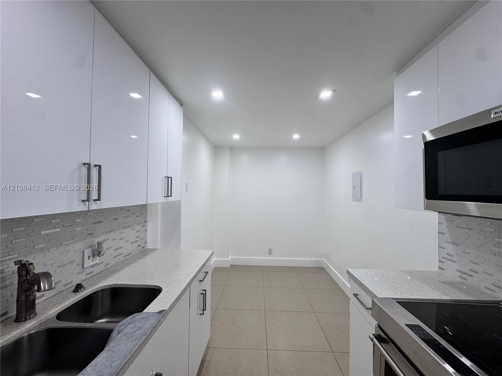 Galley kitchen featuring high-gloss white cabinetry, speckled quartz countertops, a stone mosaic tile backsplash, stainless steel appliances, and recessed lighting
