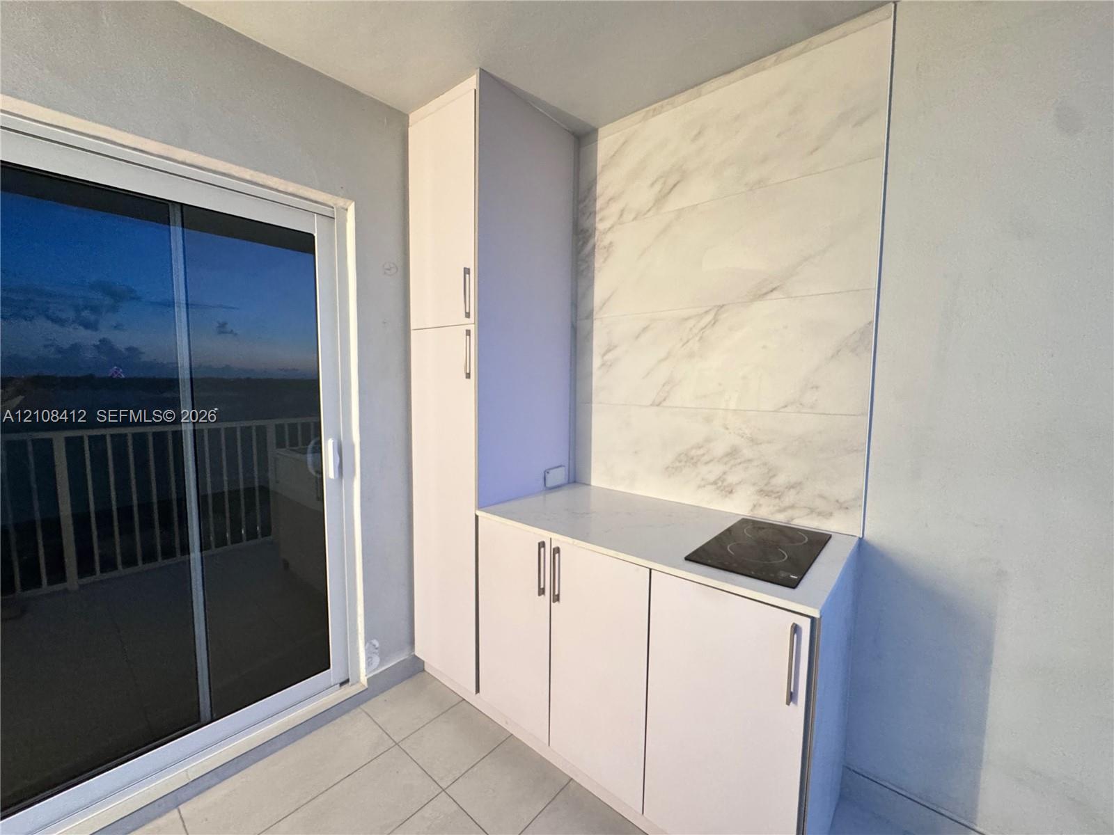 Built-in cabinetry featuring white flat-panel doors with metallic hardware, stone-finish backsplash, smooth-surface electric cooktop, and large format floor tile