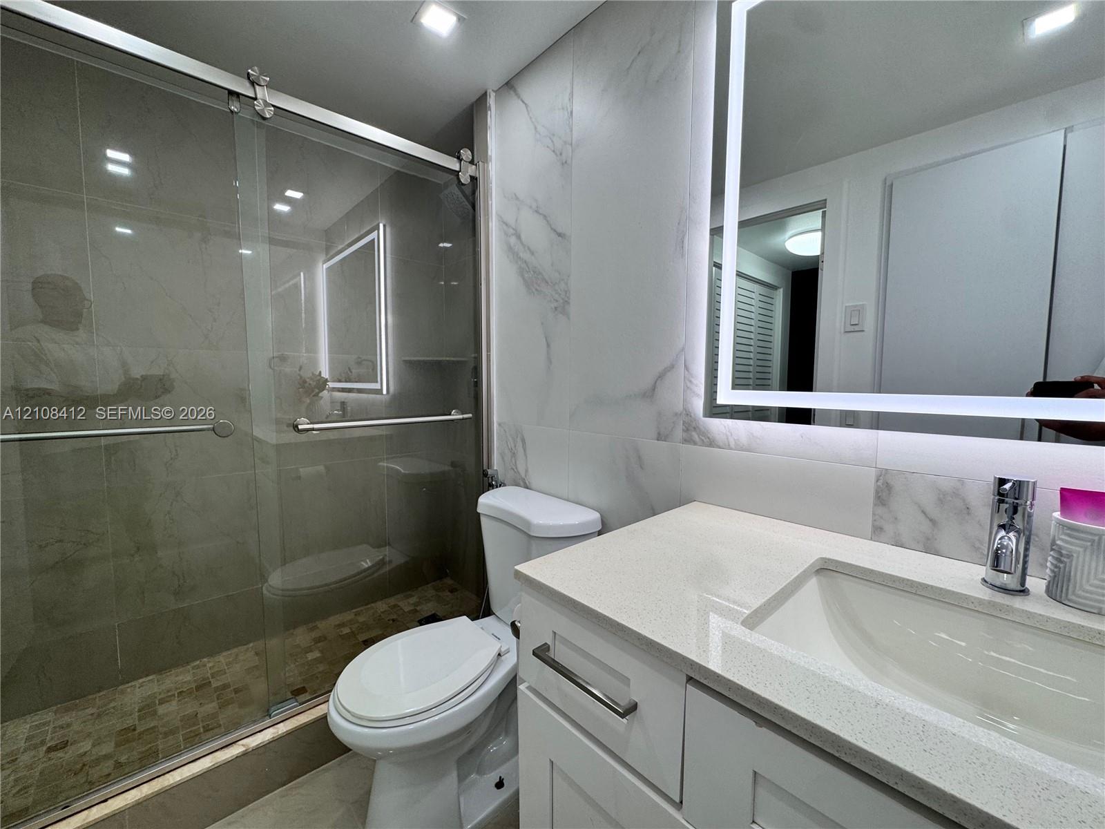 Bathroom featuring a glass-enclosed shower with tiled flooring, stone-surfaced vanity with an integrated sink, and an illuminated mirror