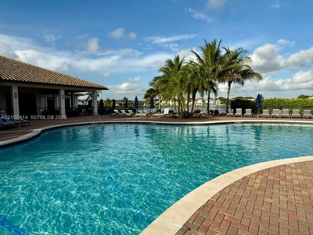 Curved swimming pool featuring a brick paver deck, covered lanai, and mature palm trees