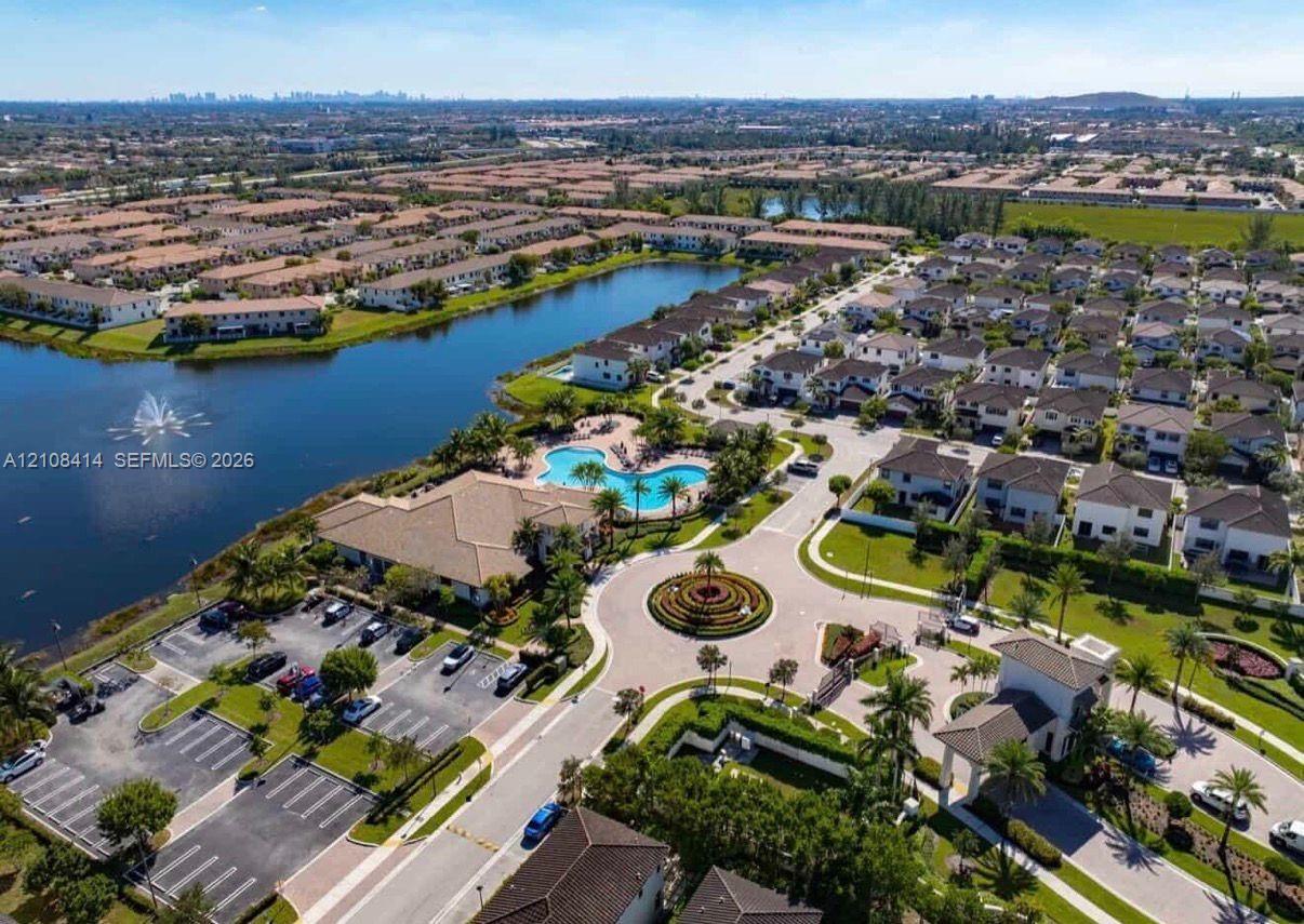 Aerial perspective of a community featuring a large retention pond with a central fountain, a swimming pool, a landscaped circular roundabout, and paved parking areas