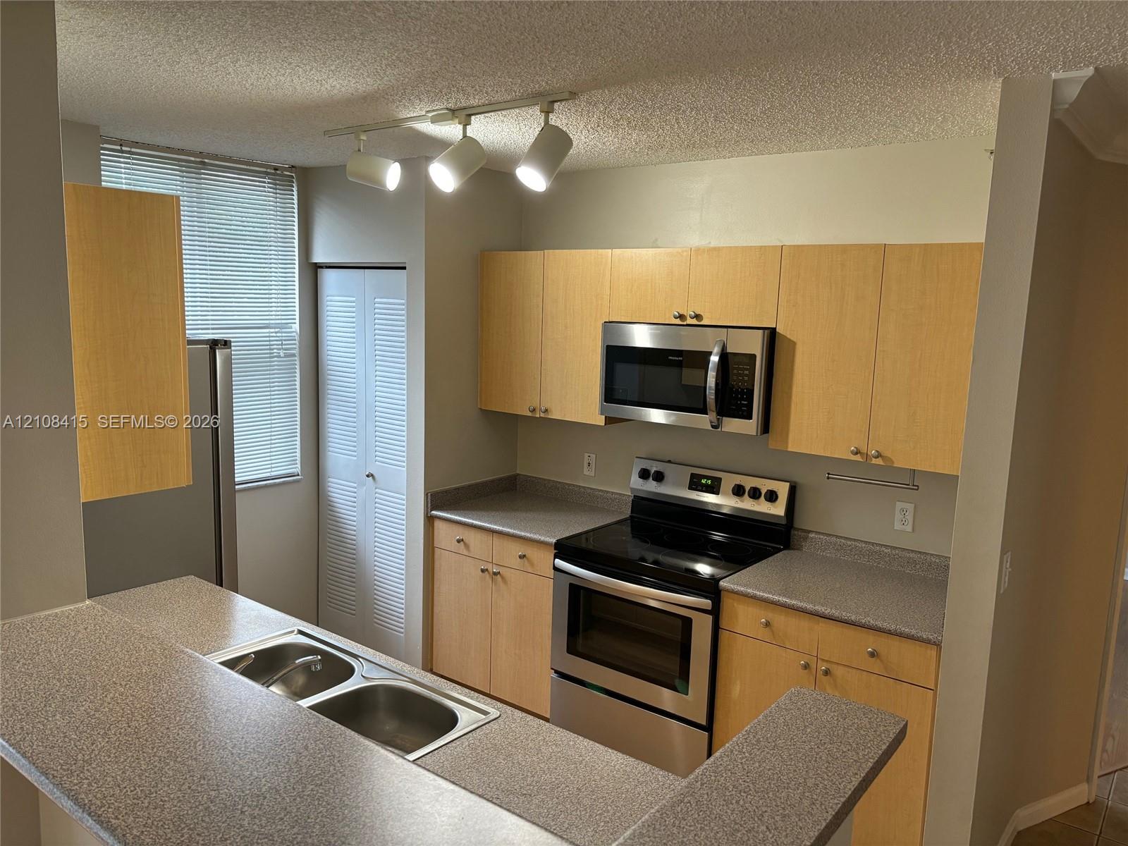 Kitchen featuring light-tone flat-panel cabinetry, stone-composite countertops, stainless steel appliances, double-basin drop-in sink, and adjustable track lighting