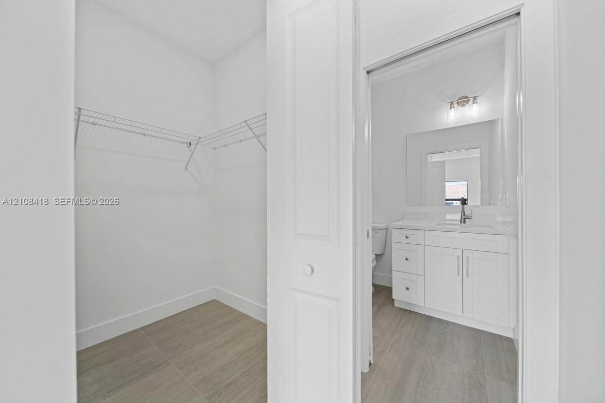 Walk-in closet featuring tiered wire shelving and wood-finish flooring, opening to a bathroom with a white vanity, quartz-style countertop, rectangular undermount sink, and wall-mounted vanity lighting