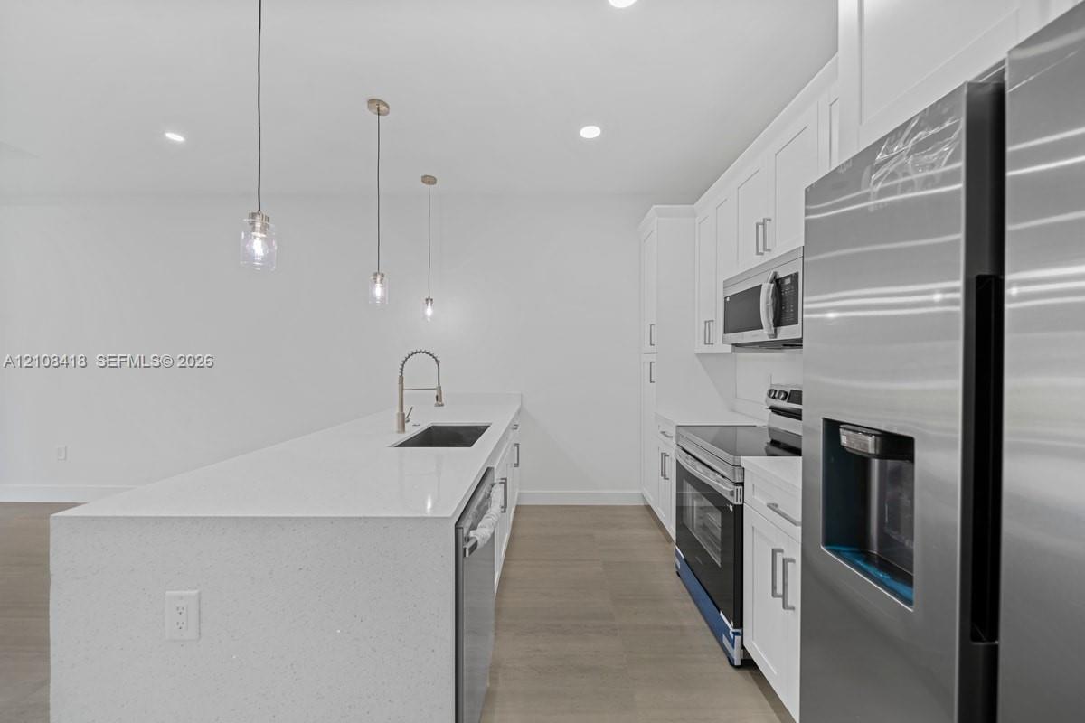 Kitchen featuring white shaker-style cabinetry, quartz countertops, an island with an undermount sink, stainless steel appliances, and wood-finish flooring
