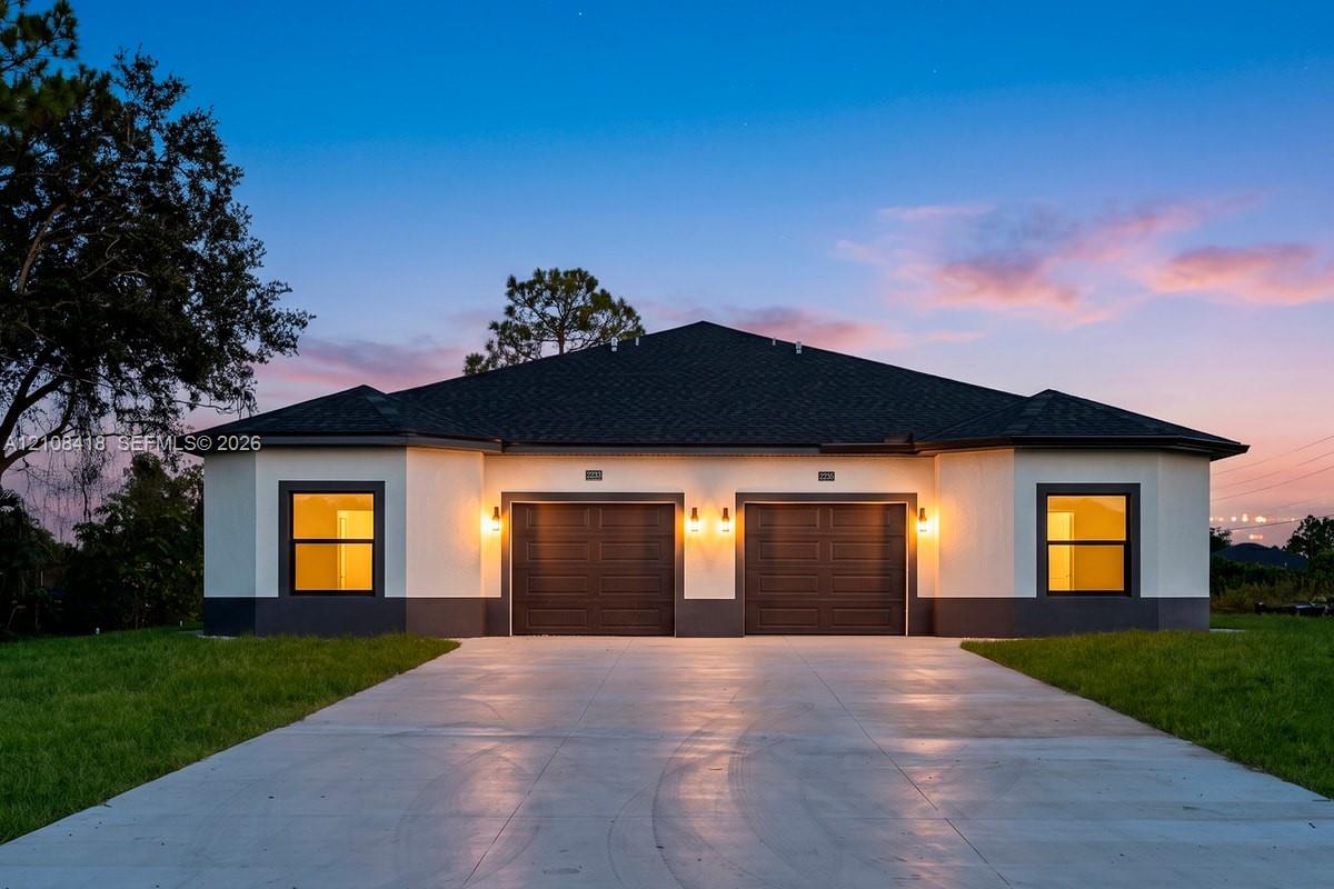 Single-story exterior featuring a light-toned stucco facade with dark contrast trim, a dual-bay garage, dark shingle roof, and a concrete driveway