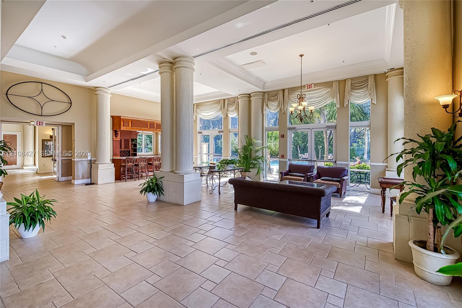 Open-concept common area featuring ceramic tile flooring, soaring tray ceilings, structural columns, built-in wooden bar cabinetry, and floor-to-ceiling glass panel doors