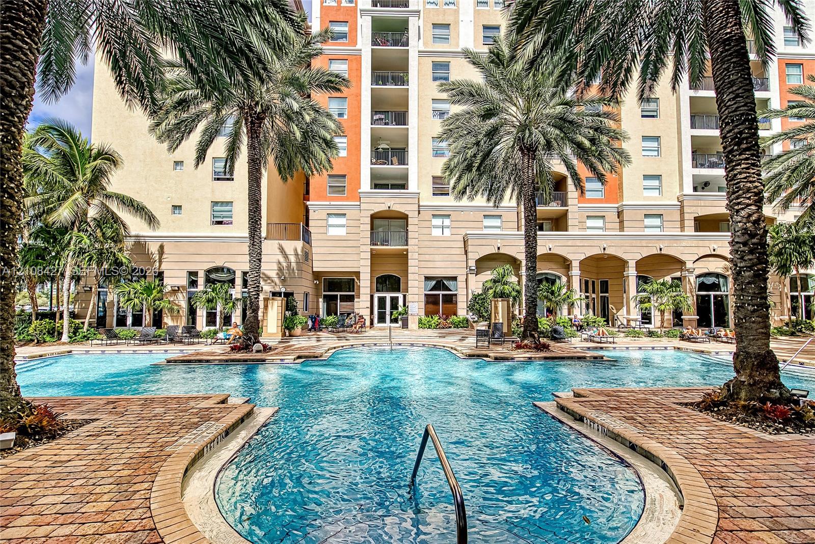 Resort-style swimming pool featuring a brick-paver deck, arched architectural detailing, mature palm trees, and multi-story residential facade Resort-style swimming pool featuring a brick-paver deck, arched architectural detailing, mature palm trees, and multi-story residential facade