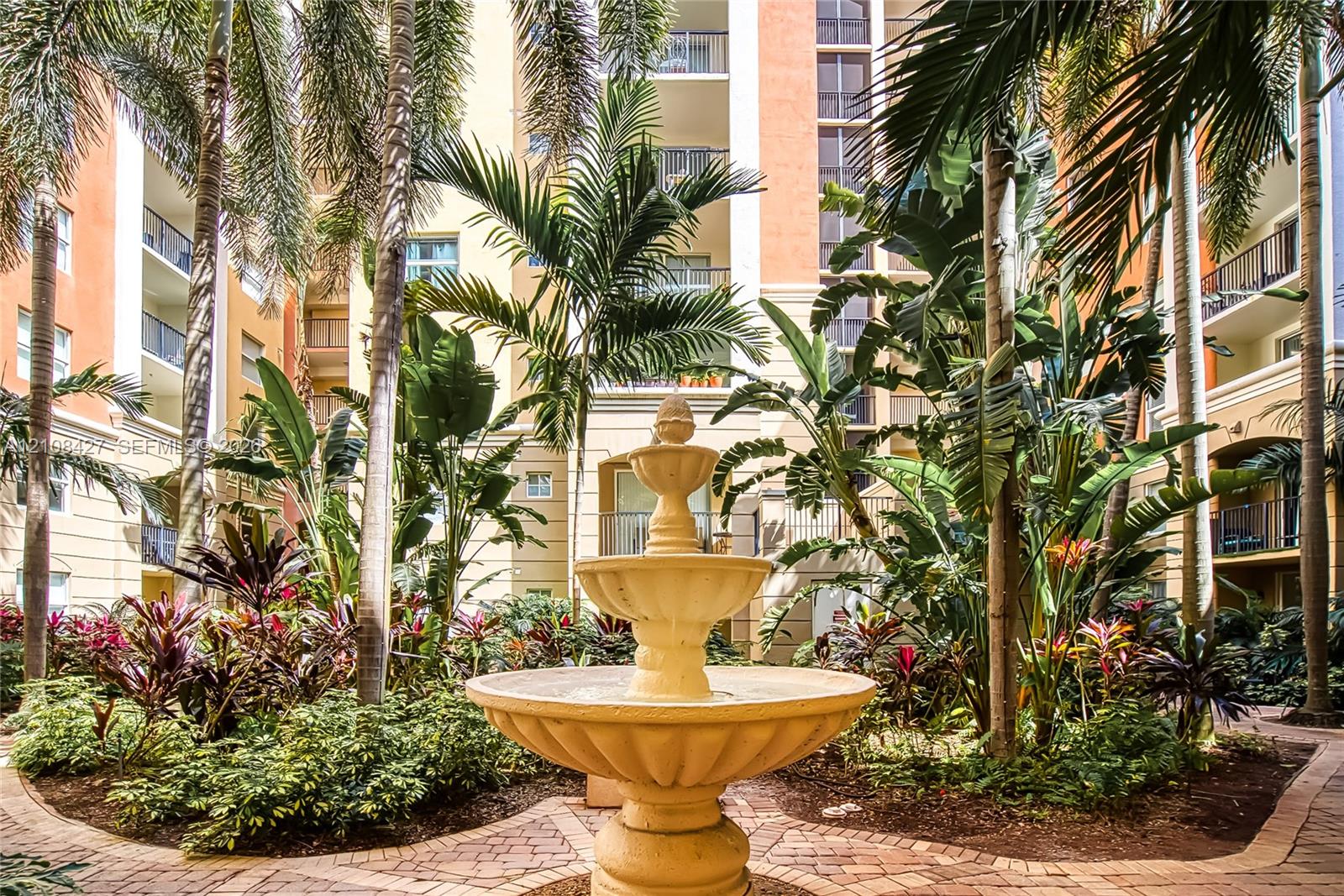Central courtyard featuring a multi-tiered stone fountain, paved brick pathways, mature palm trees, and lush tropical landscaping