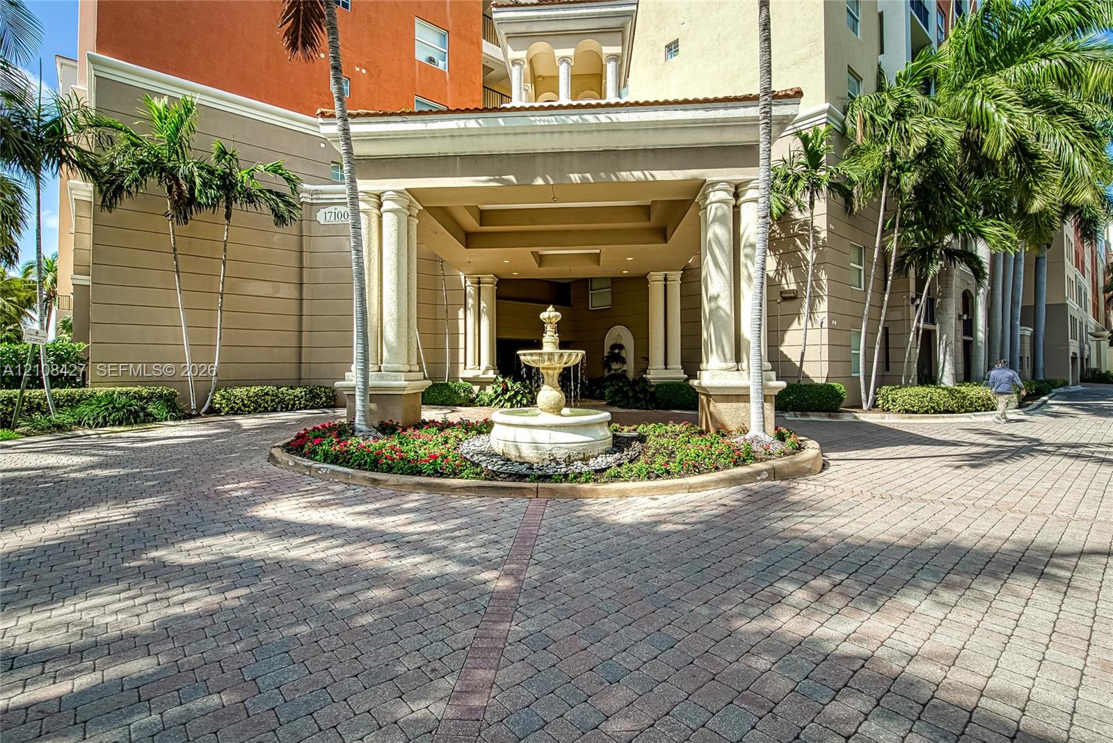 Covered motor court entrance featuring a tiered stone fountain, paved circular driveway, architectural columns, and tropical landscaping