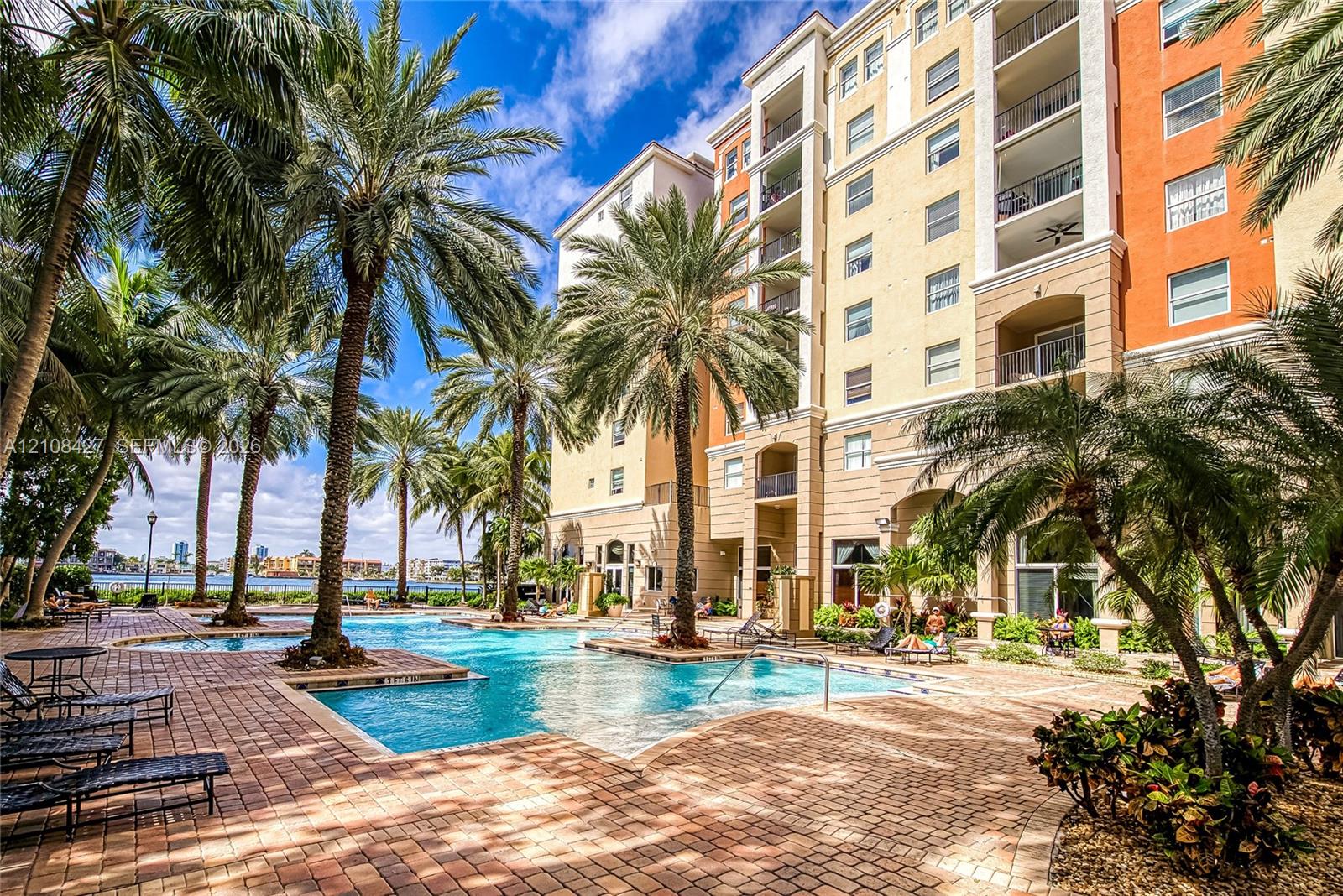 Multi-story stucco exterior featuring private balconies and an expansive brick-paver pool deck surrounded by mature palm trees