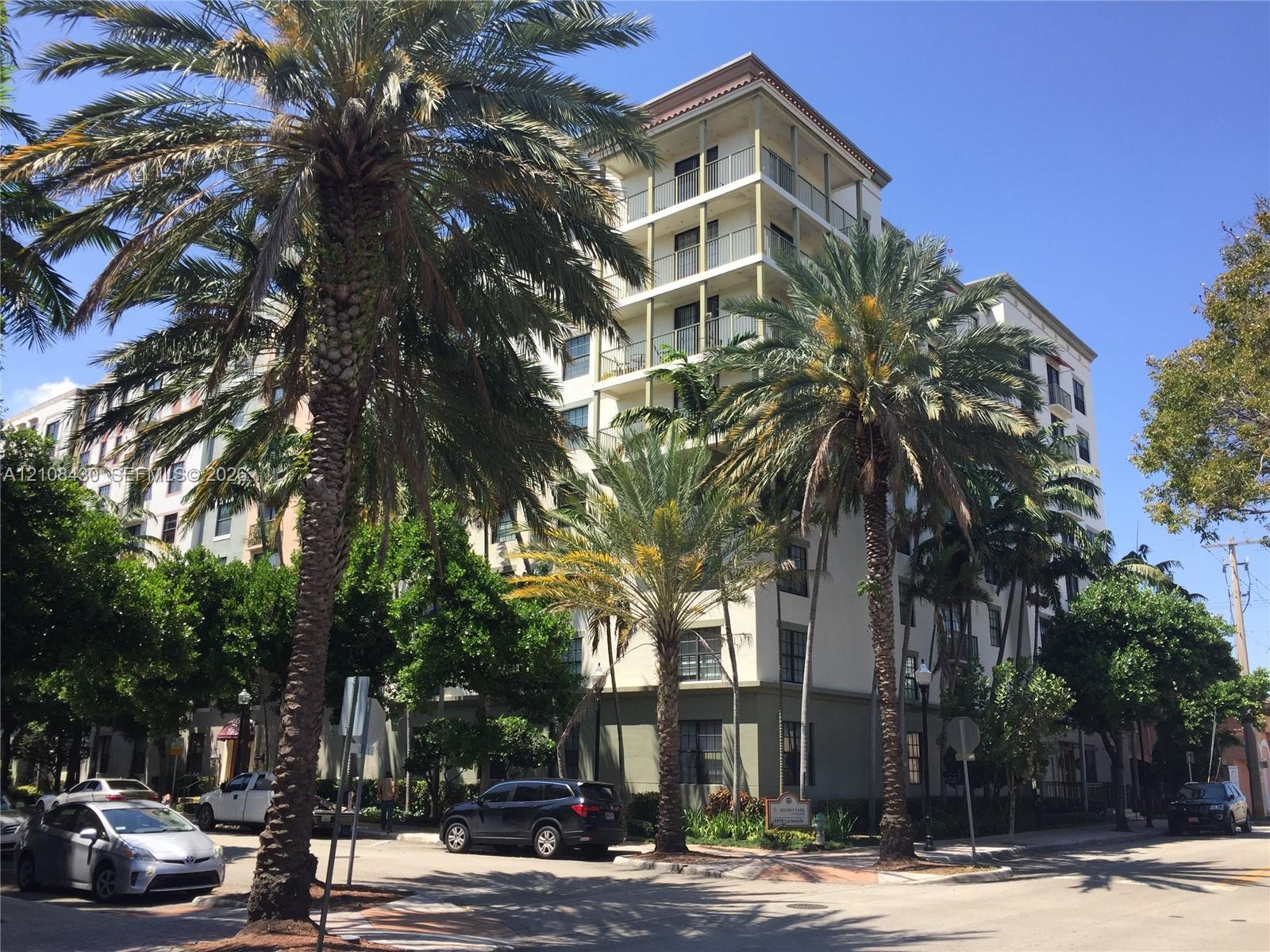 Mid-rise multi-story building exterior featuring a stucco facade, tiered balconies with glass railings, a flat roofline, and mature palm tree landscaping
