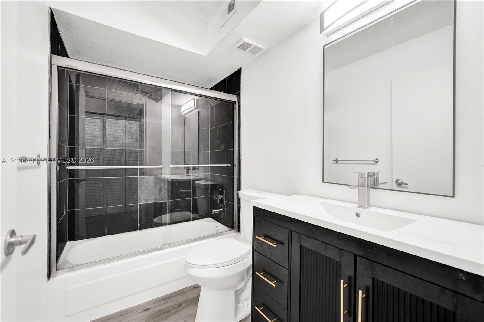 Bathroom featuring a modern vanity with a solid-surface countertop, an integrated rectangular sink, black cabinetry with gold-tone hardware, a large frameless mirror, and a shower-tub combination with dark tile surround