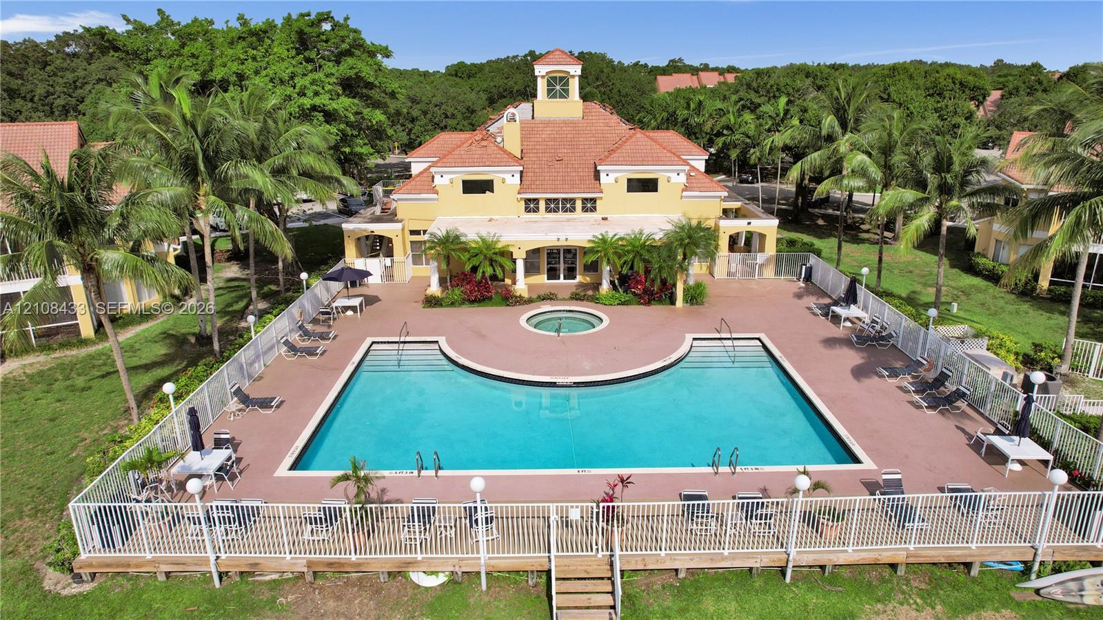 Mediterranean-style clubhouse featuring a terra-cotta tile roof and a central observation tower, accompanied by a fenced outdoor pool and spa surrounded by a paved deck