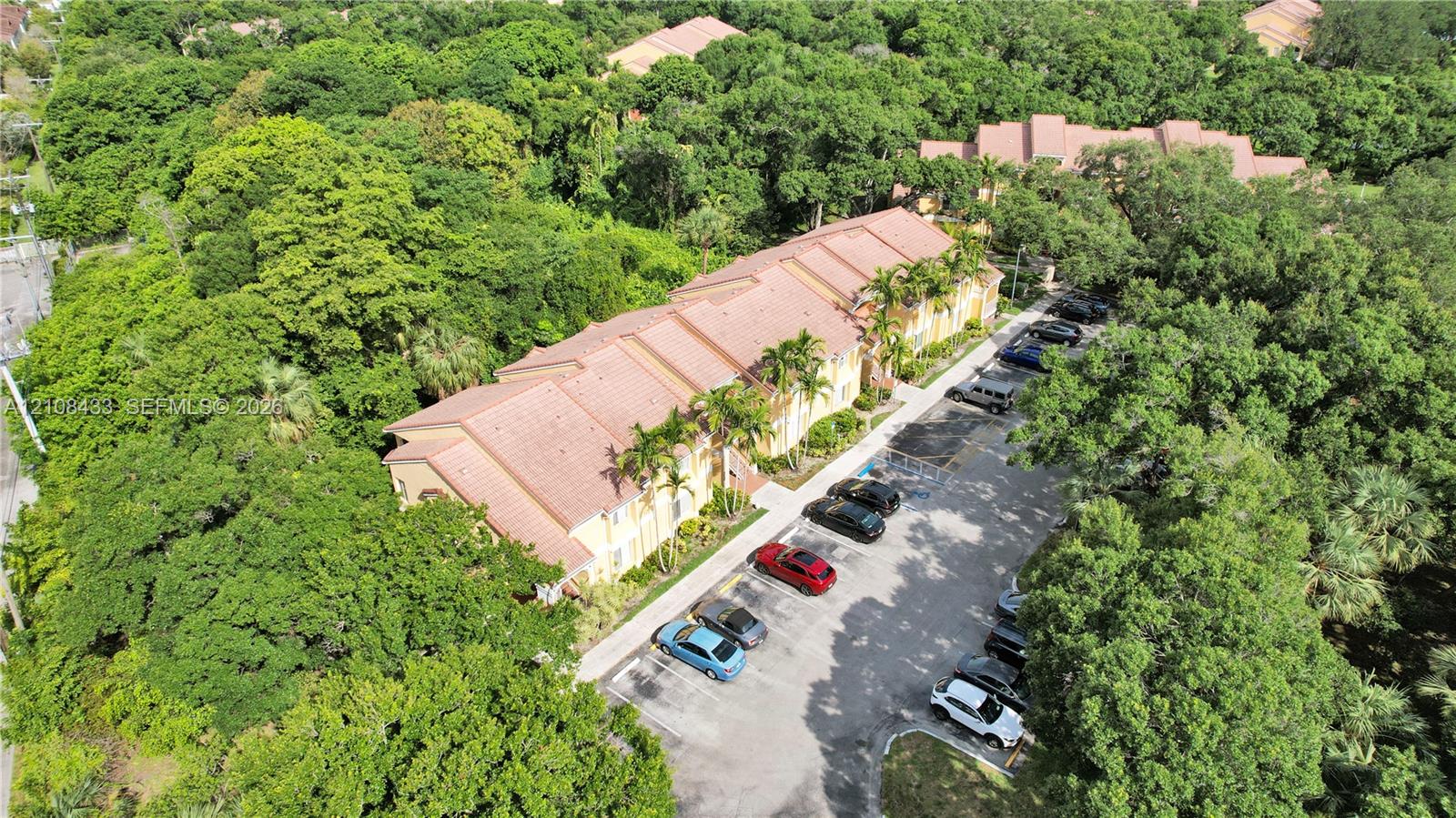 Multi-story building featuring a tile roof, exterior stucco siding, and designated asphalt surface parking, situated among mature trees and dense vegetation Multi-story building featuring a tile roof, exterior stucco siding, and designated asphalt surface parking, situated among mature trees and dense vegetation