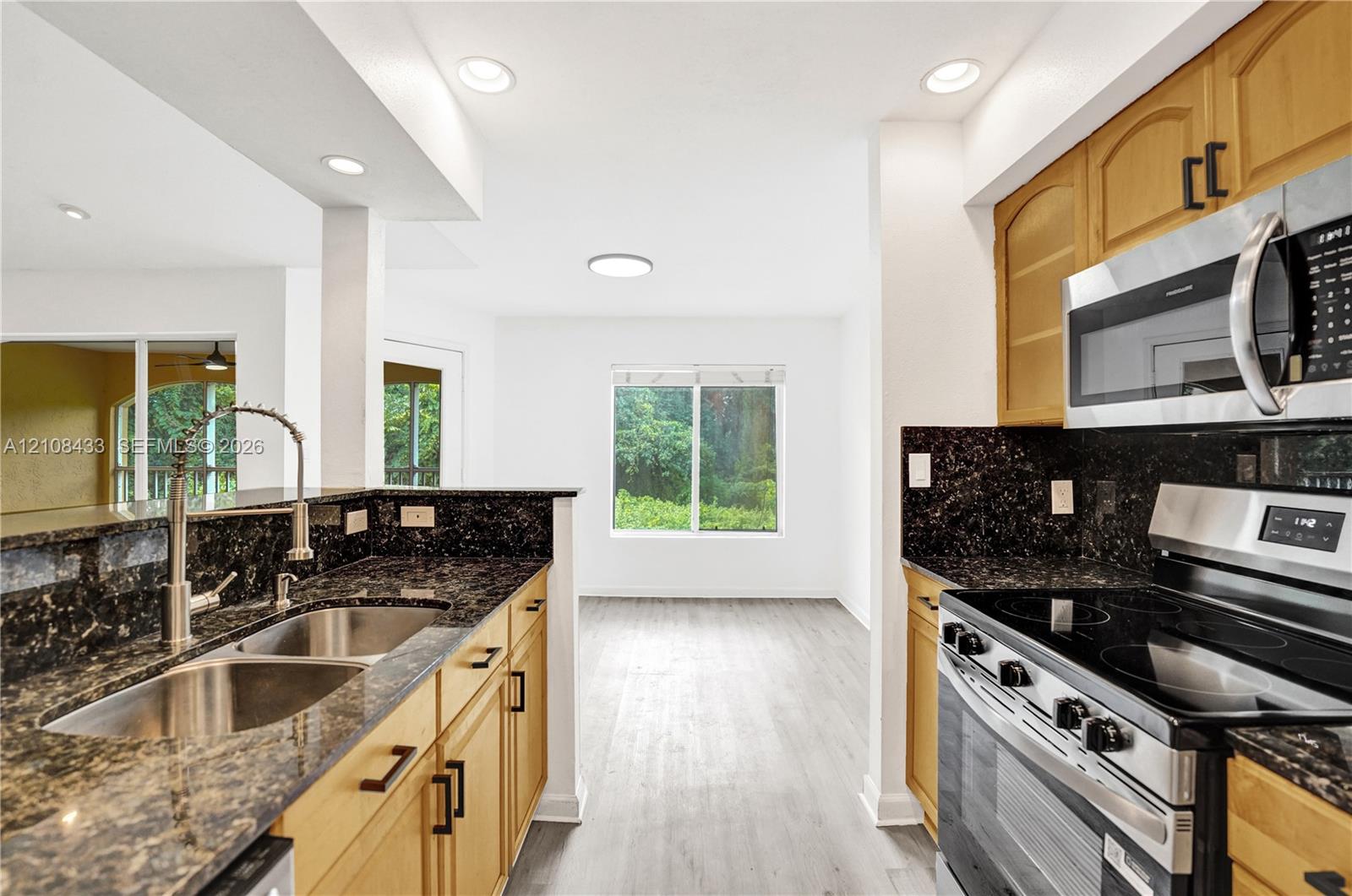 Kitchen featuring medium-tone wood cabinetry, dark stone countertops, stainless steel appliances, a dual-basin undermount sink, and wood-finish flooring Kitchen featuring medium-tone wood cabinetry, dark stone countertops, stainless steel appliances, a dual-basin undermount sink, and wood-finish flooring